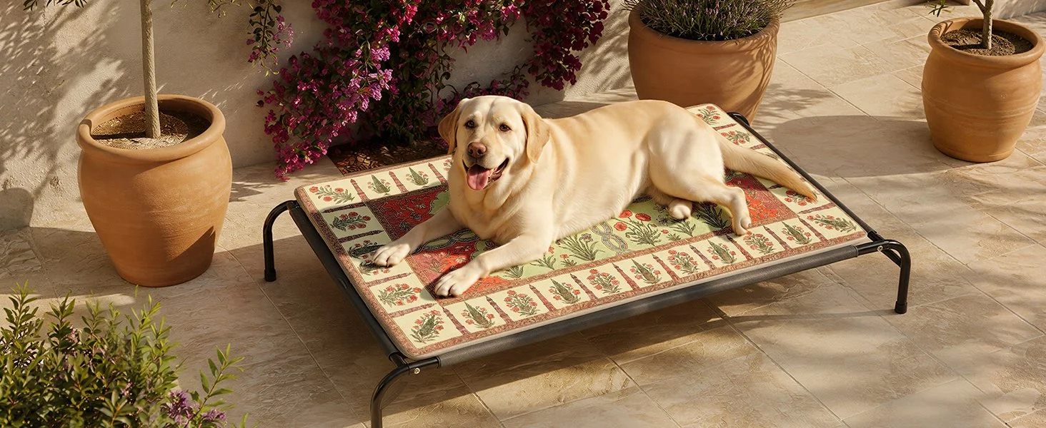 A happy yellow Labrador Retriever lying on a cushioned outdoor pet bed with a floral and Christmas-themed pattern, surrounded by potted plants and flowering bushes on a tiled patio.