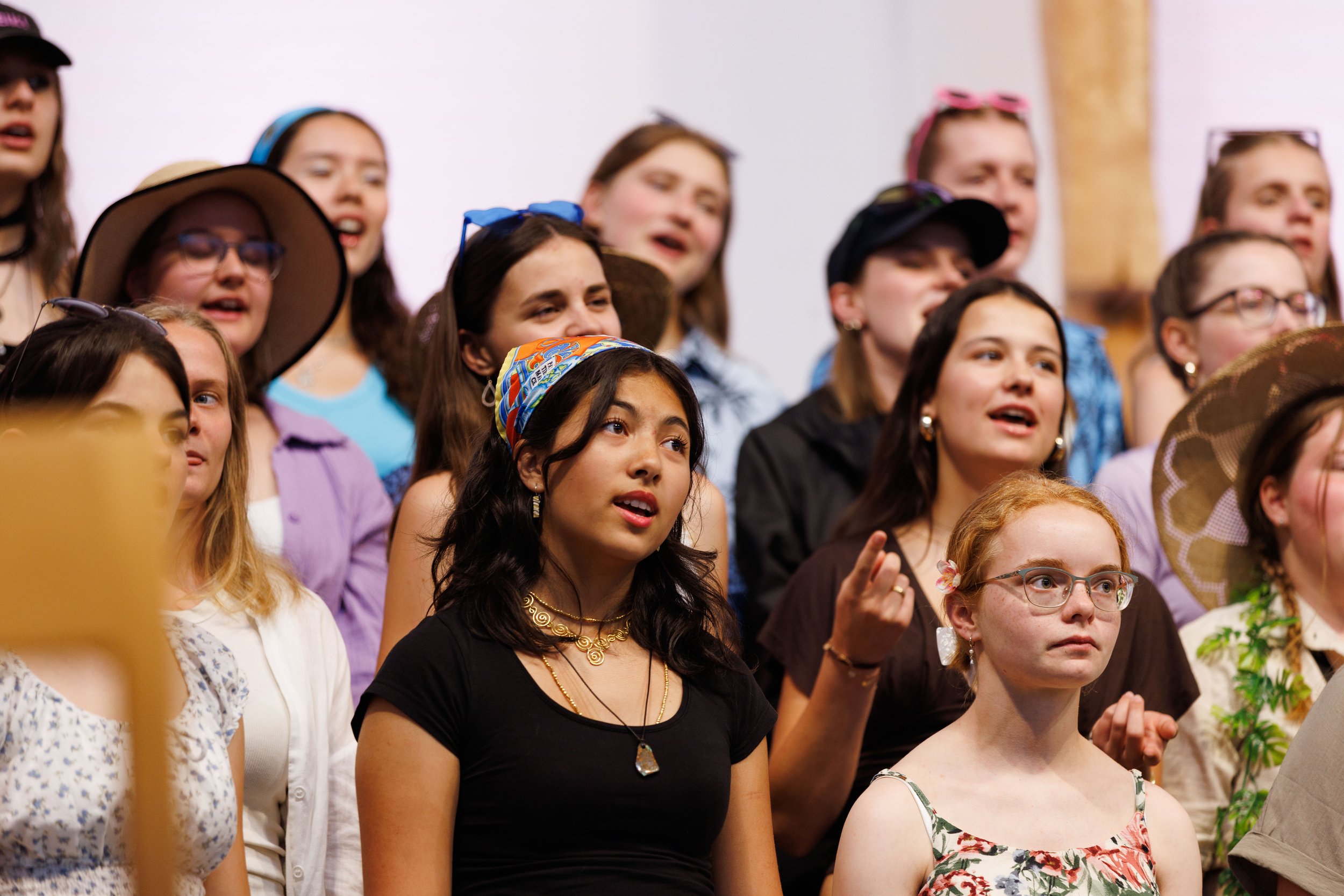 Eine Gruppe junger Frauen und Mädchen singt oder tritt auf, einige tragen Hüte und Sonnenbrillen, in einem Innenraum mit weißem Hintergrund.