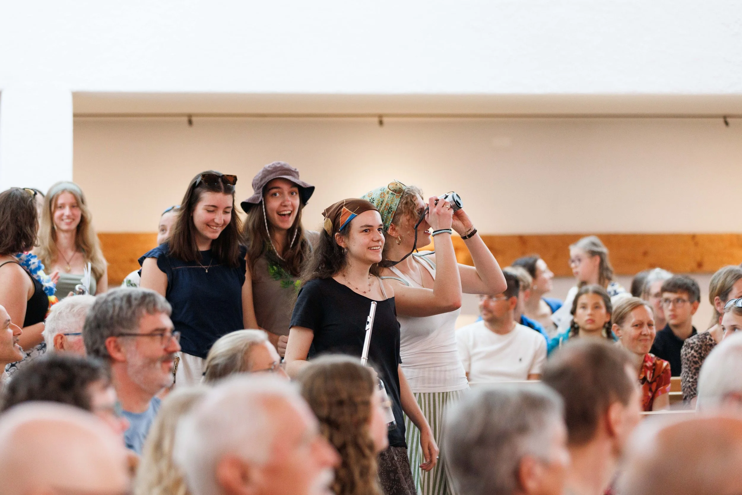Jugendliche und Erwachsene versammeln sich in einem Saal, einige stehen, andere sitzen, einige fotografieren oder schauen interessiert. Es ist eine lebhafte Veranstaltung.