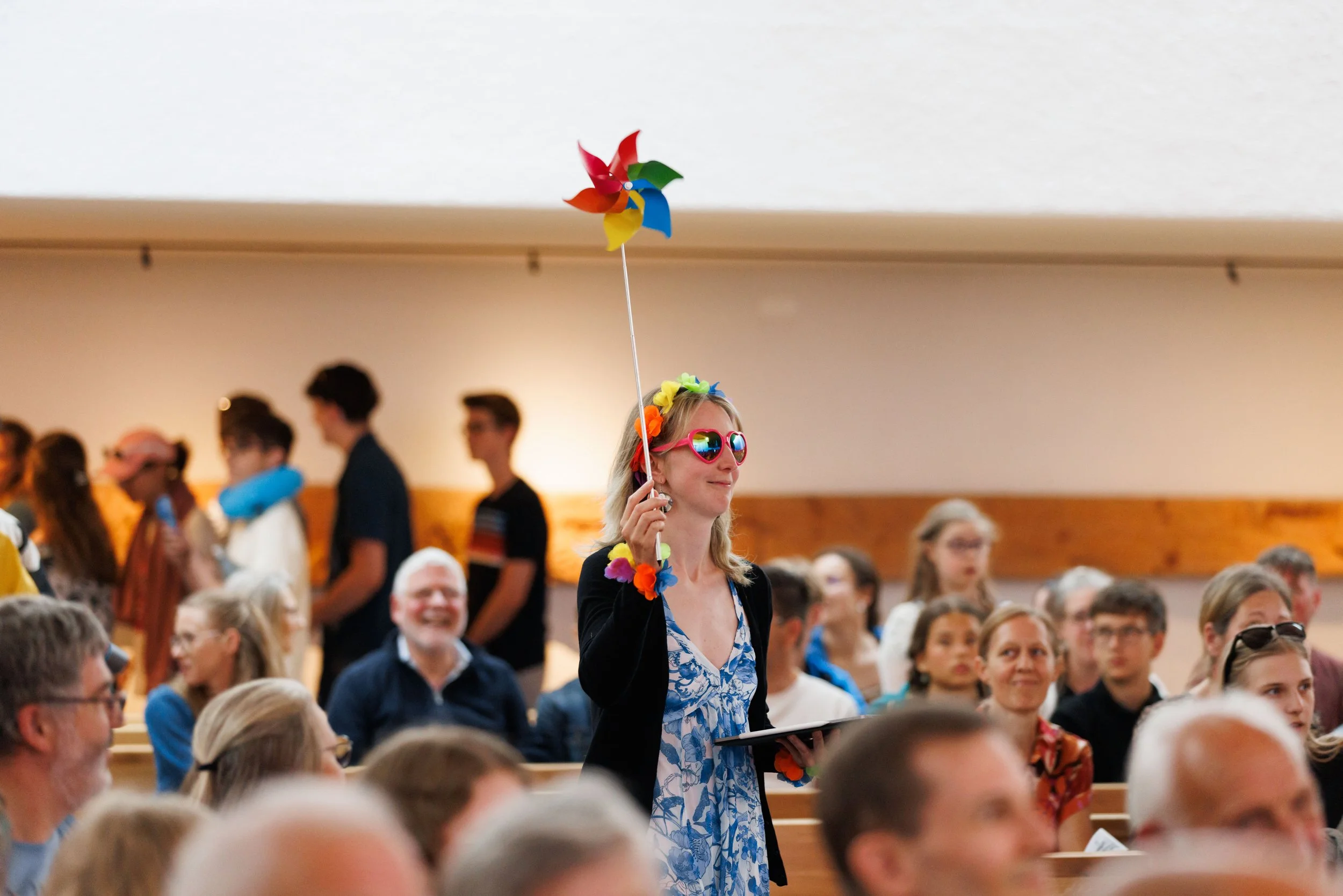 Frau mit buntem Blumenkranz und Sonnenbrille hält ein buntes Windrad in einer Menschenmenge bei einer Veranstaltung.