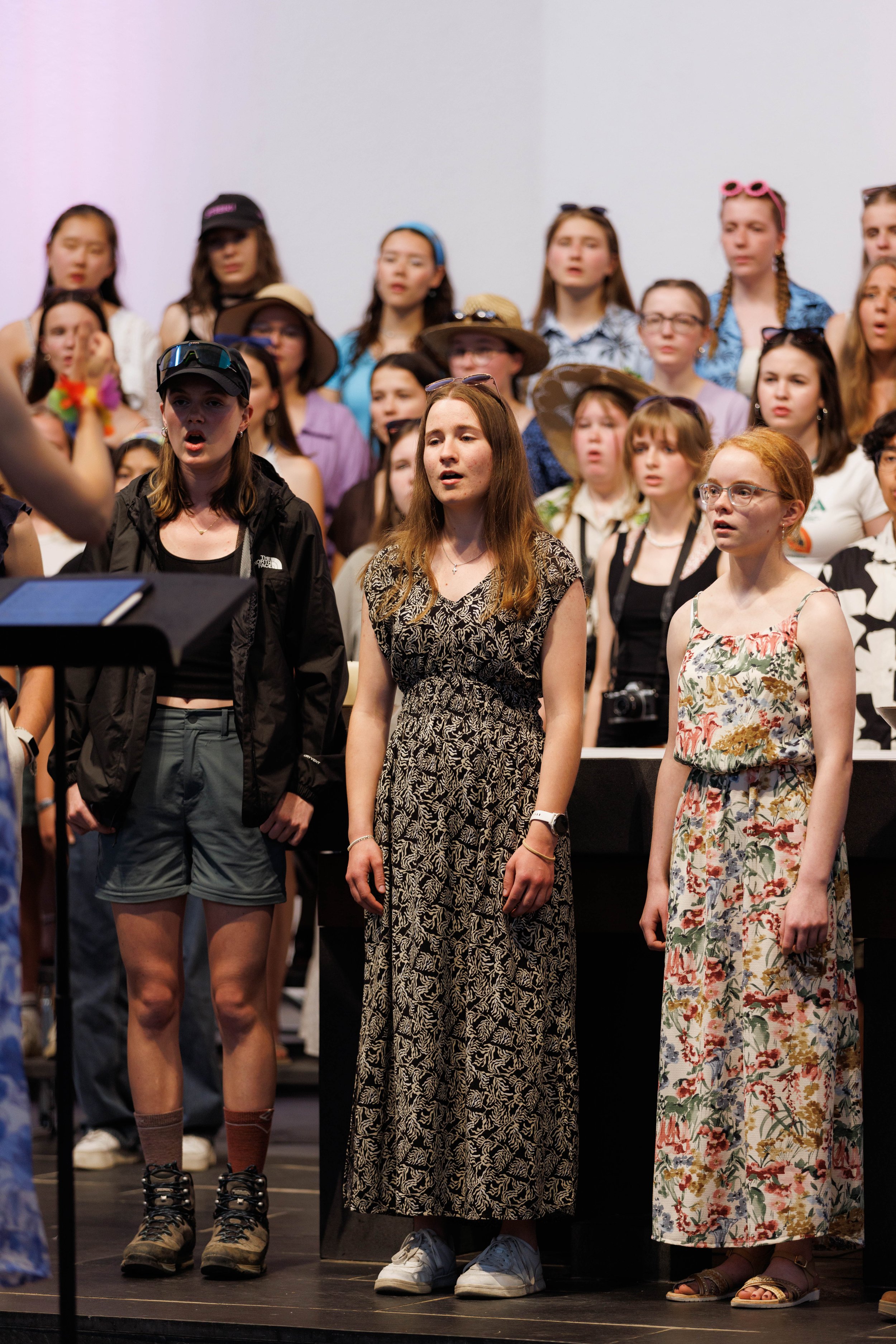 Eine Gruppe junger Frauen singt auf einer Bühne, einige tragen Hüte oder Brillen, während sie sich auf einen Auftritt vorbereiten.