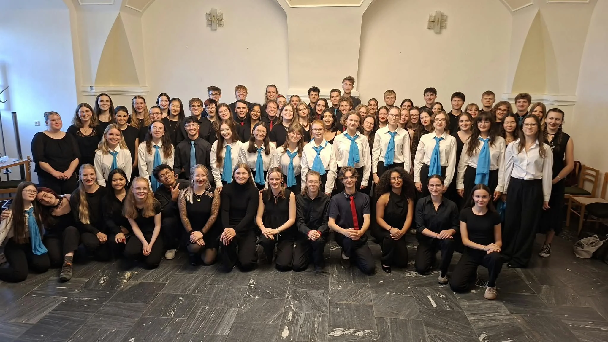 Gruppe von Jugendlichen in formeller Kleidung, einige mit blauen Halstüchern, bei einer Veranstaltung in einem Saal