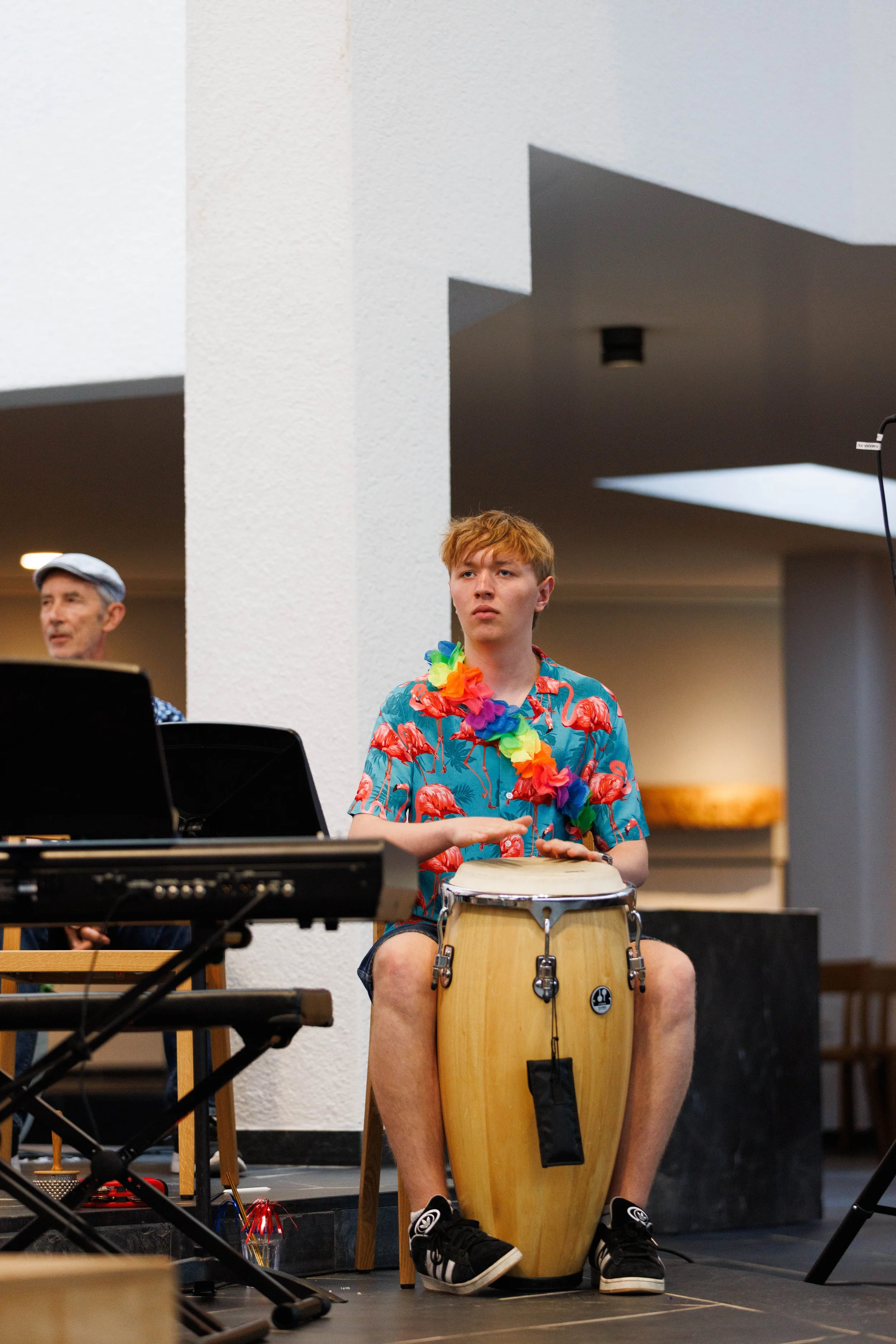 Junge Person mit roten Haaren sitzt auf einem Stuhl und spielt Conga-Trommel, trägt ein buntes Hawaihemd mit Flamingos und eine bunte Halskette. Im Hintergrund sind weitere Musiker und Musikinstrumente, die Szene wirkt wie eine musikalische Darbietun
