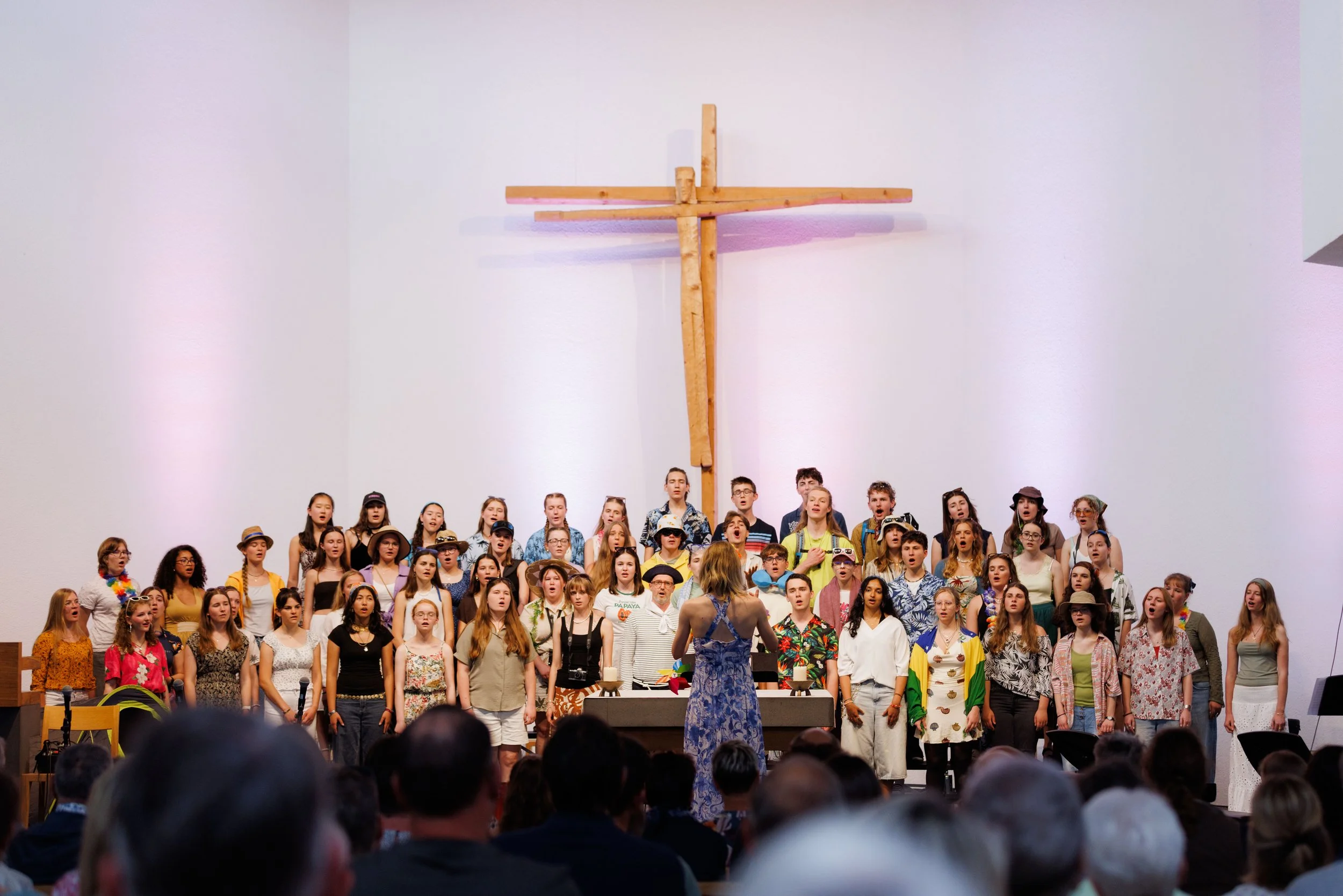 Chor von Jugendlichen und Erwachsenen, die auf einer Bühne singen, unter Leitung einer Dirigentin, mit einem großen Kreuz im Hintergrund vor einer weißen Wand.
