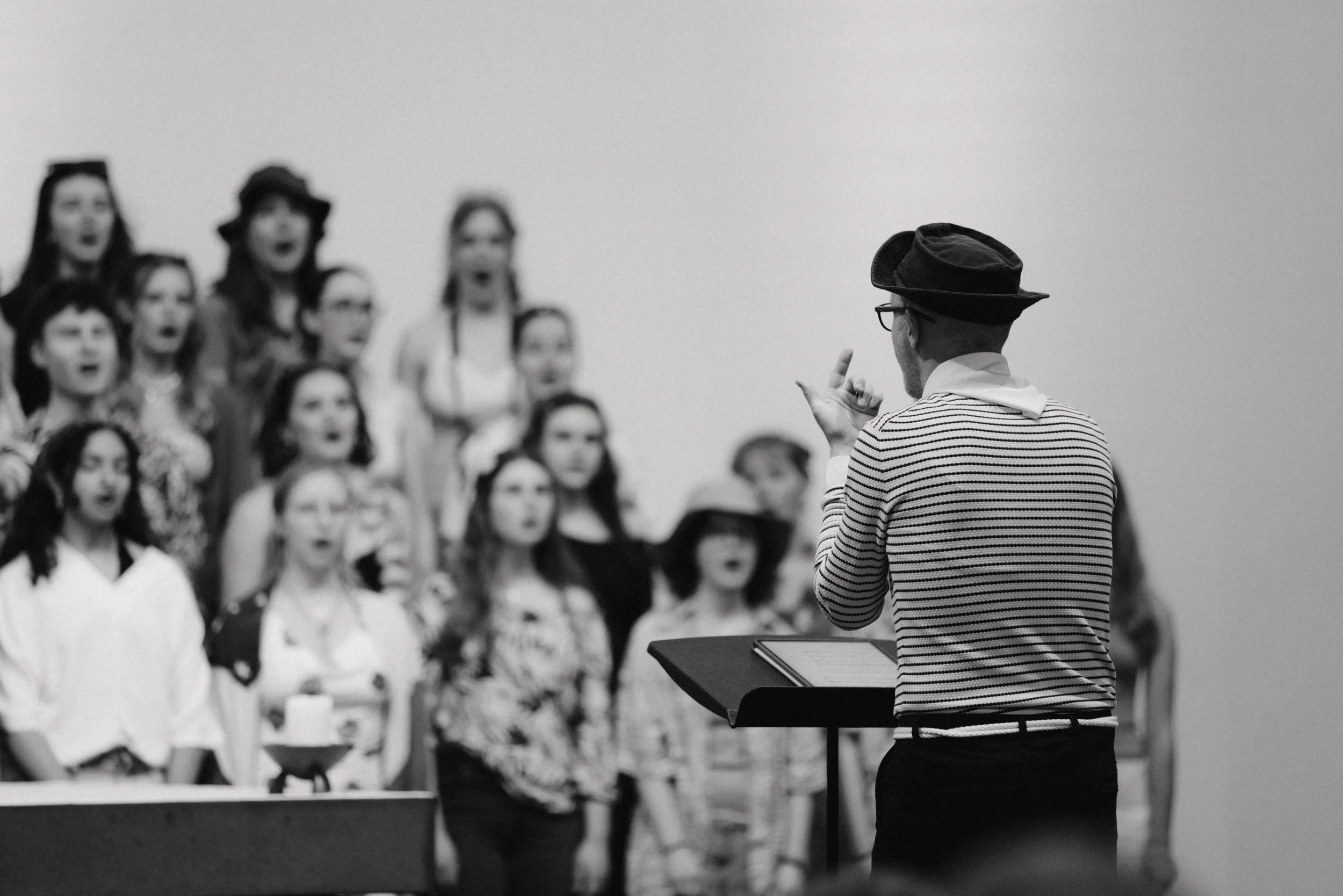 Ein Chor von Frauen singt, während ein Mann mit Hut und gestreiftem Hemd vor ihnen steht, vermutlich Dirigent, in einem Probenraum.