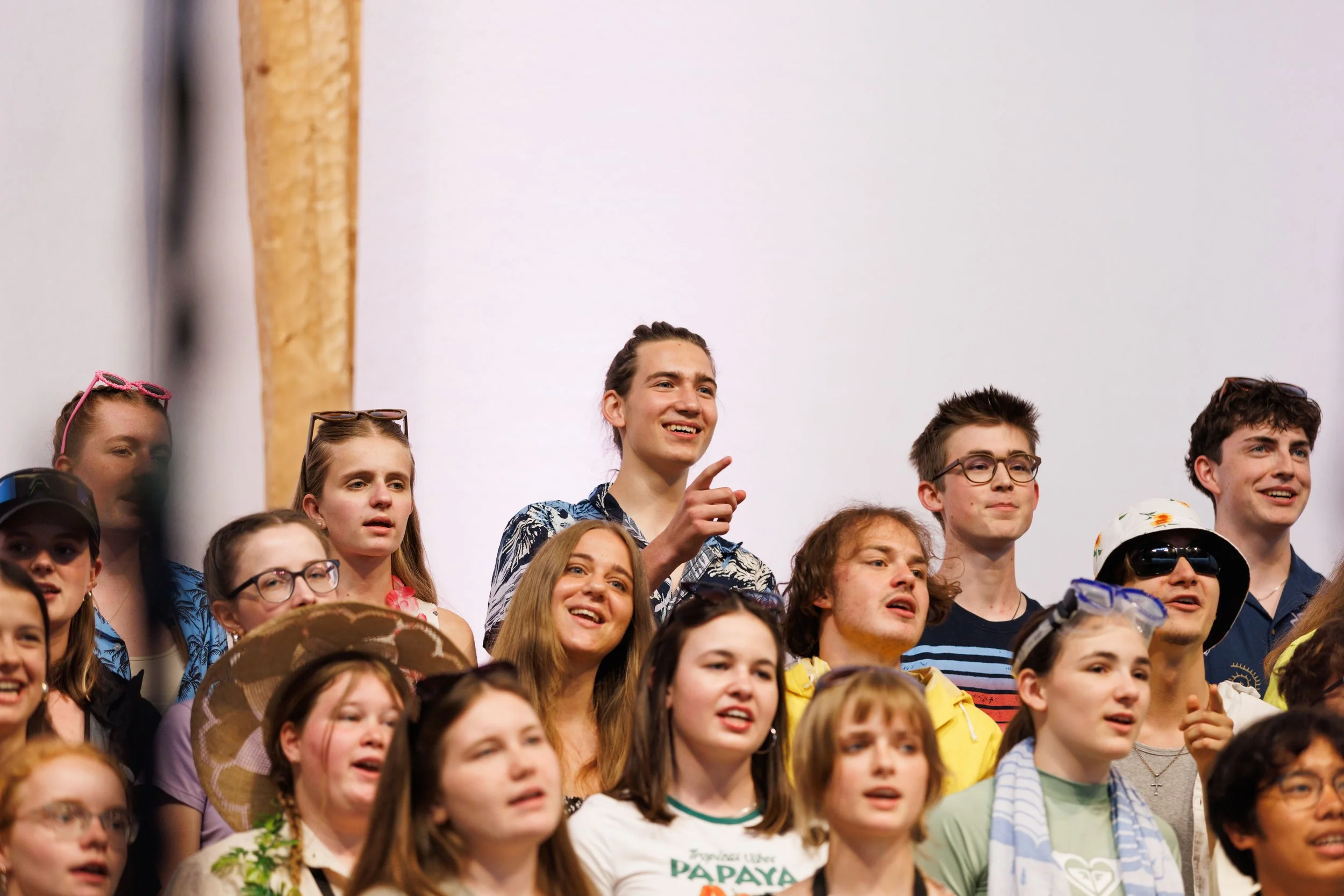 Eine große Gruppe junger Menschen, die zusammen auf einem Foto posieren, einige lachen, andere schauen in verschiedene Richtungen, tragen sommerliche Kleidung und Sonnenbrillen.