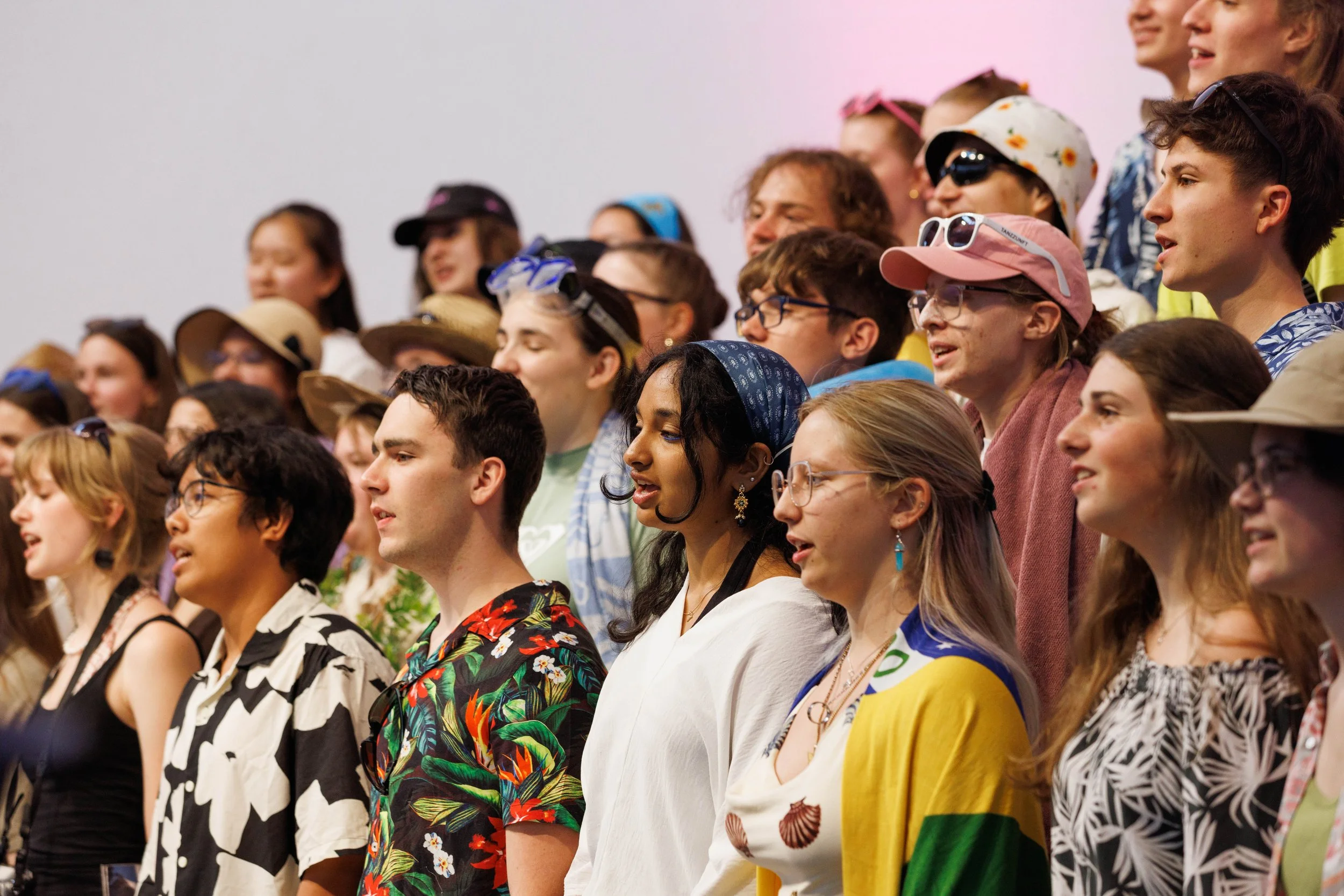 Eine Gruppe junger Menschen beim Singen oder Sprechen, viele tragen sommerliche Kleidung und Accessoires wie Sonnenbrillen und Hüte, stehen nebeneinander gegen einen weißen Hintergrund.