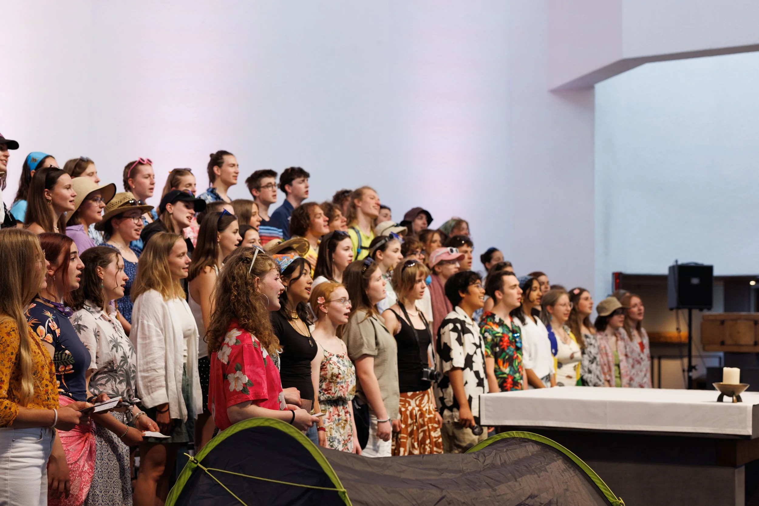 Eine Gruppe junger Menschen, überwiegend Frauen, stehen nebeneinander in einer Halle und singen oder schauen aufmerksam nach vorne, während sie an einem Event teilnehmen. Einige tragen Sonnenbrillen, Hüte und bunte Kleidung.