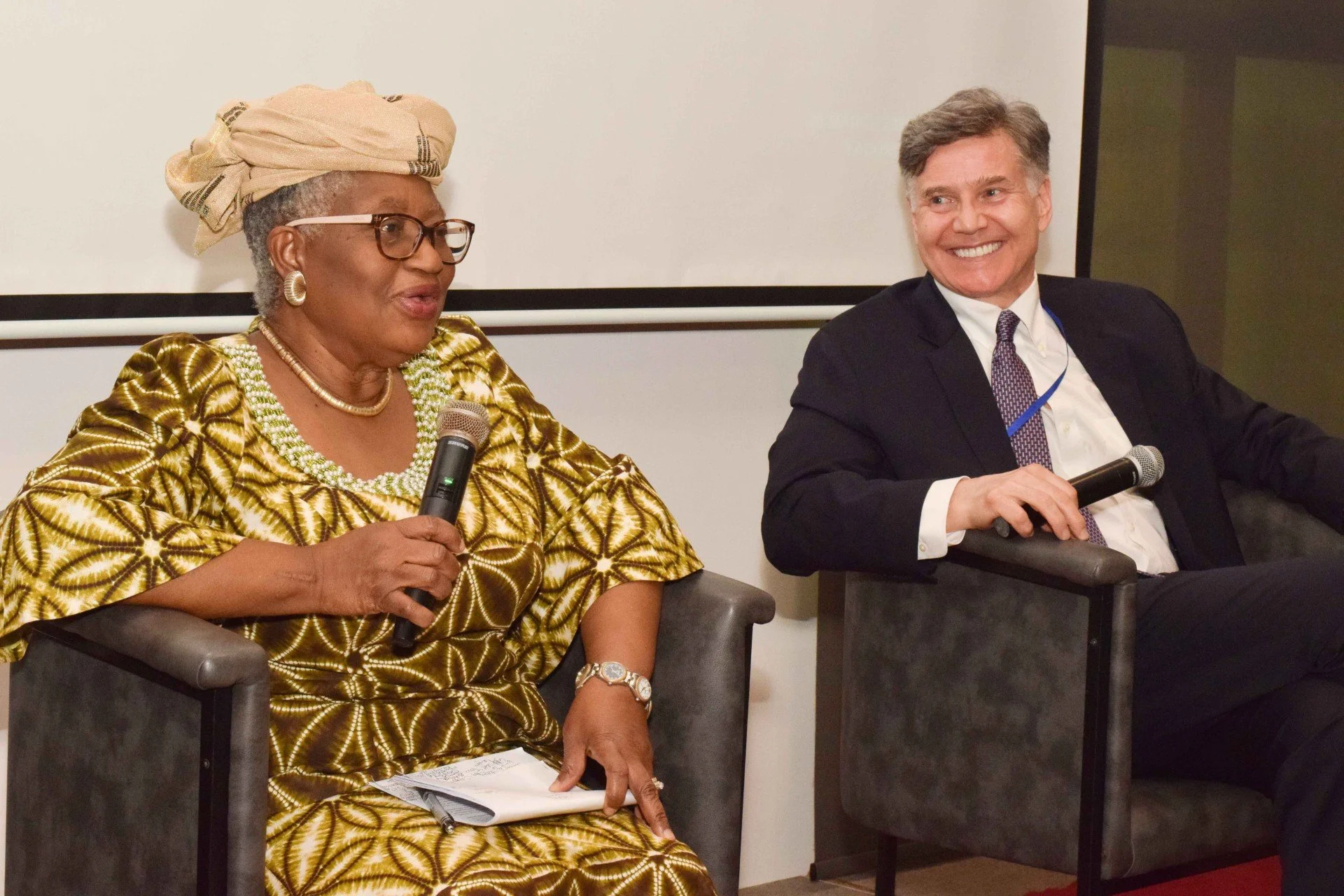 Two people sitting in chairs at a panel discussion, holding microphones. The woman on the left is wearing glasses, a headwrap, and a gold patterned dress. The man on the right is wearing a dark suit and tie, smiling.