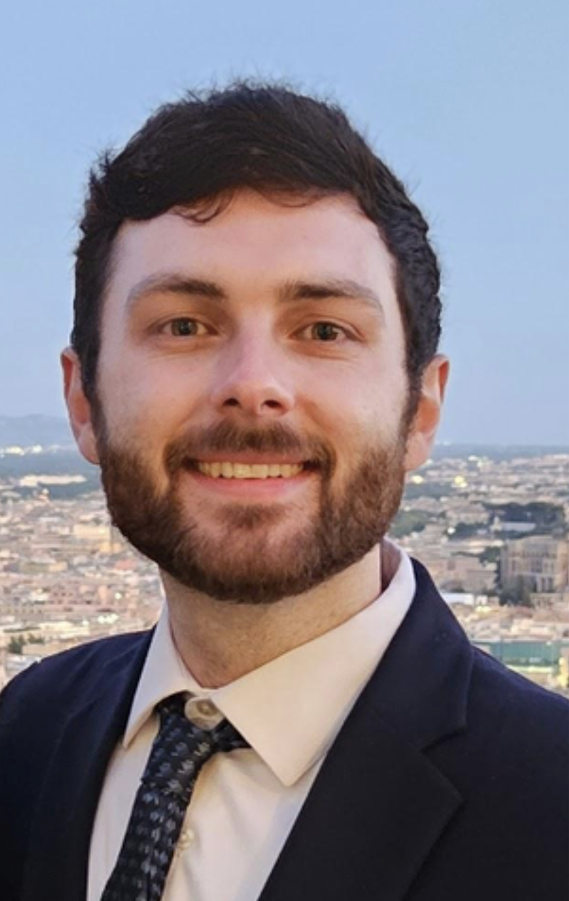 A young man with short dark hair and a beard, wearing a black suit and a patterned tie, is smiling for a photo against a cityscape background during the daytime.