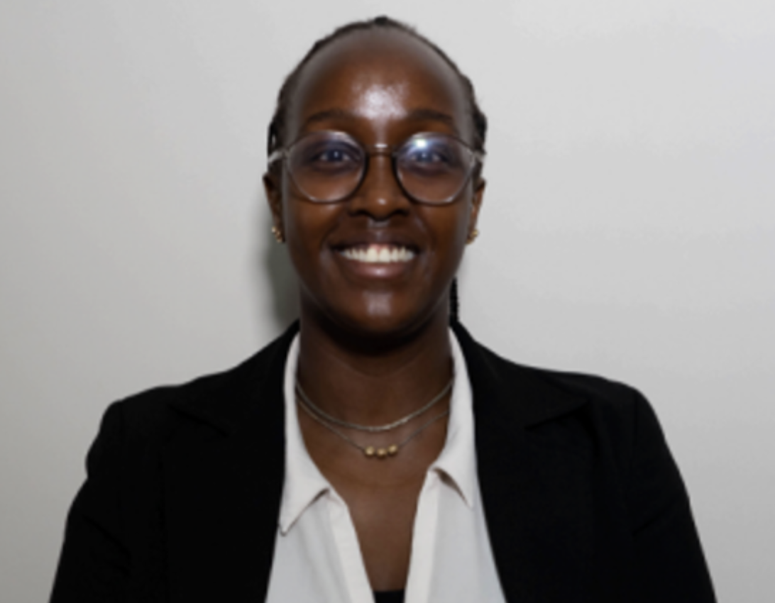 A smiling woman with dark skin, glasses, and earrings, wearing a black blazer and white shirt, standing against a plain light-colored wall.