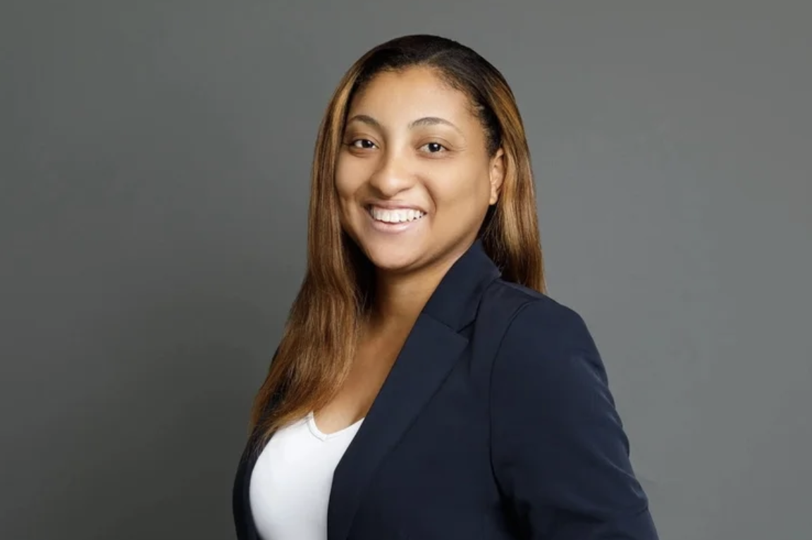 Portrait of a smiling woman with shoulder-length hair in a navy blazer and white top, standing against a gray background.