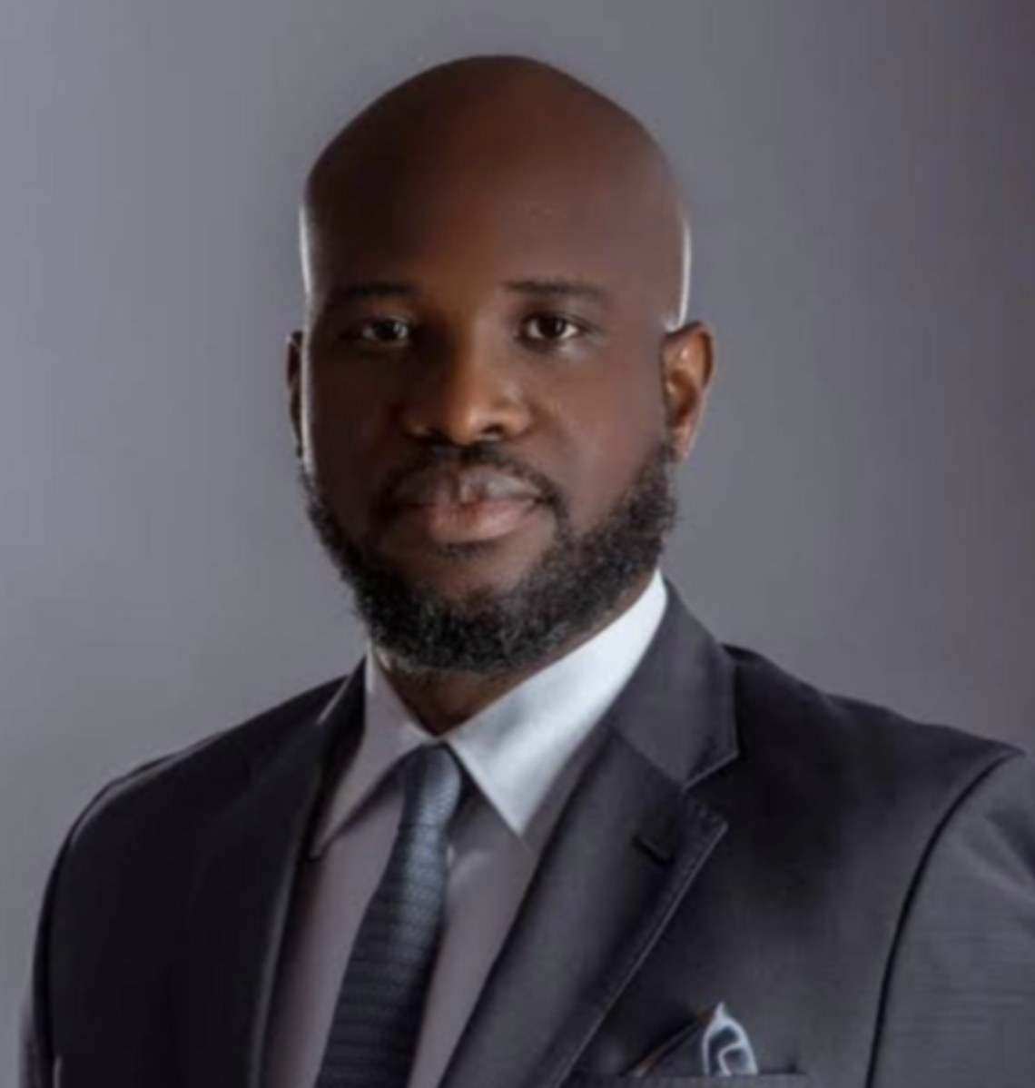 Portrait of a Black man with a beard in a dark suit, white shirt, and tie, against a gray background.