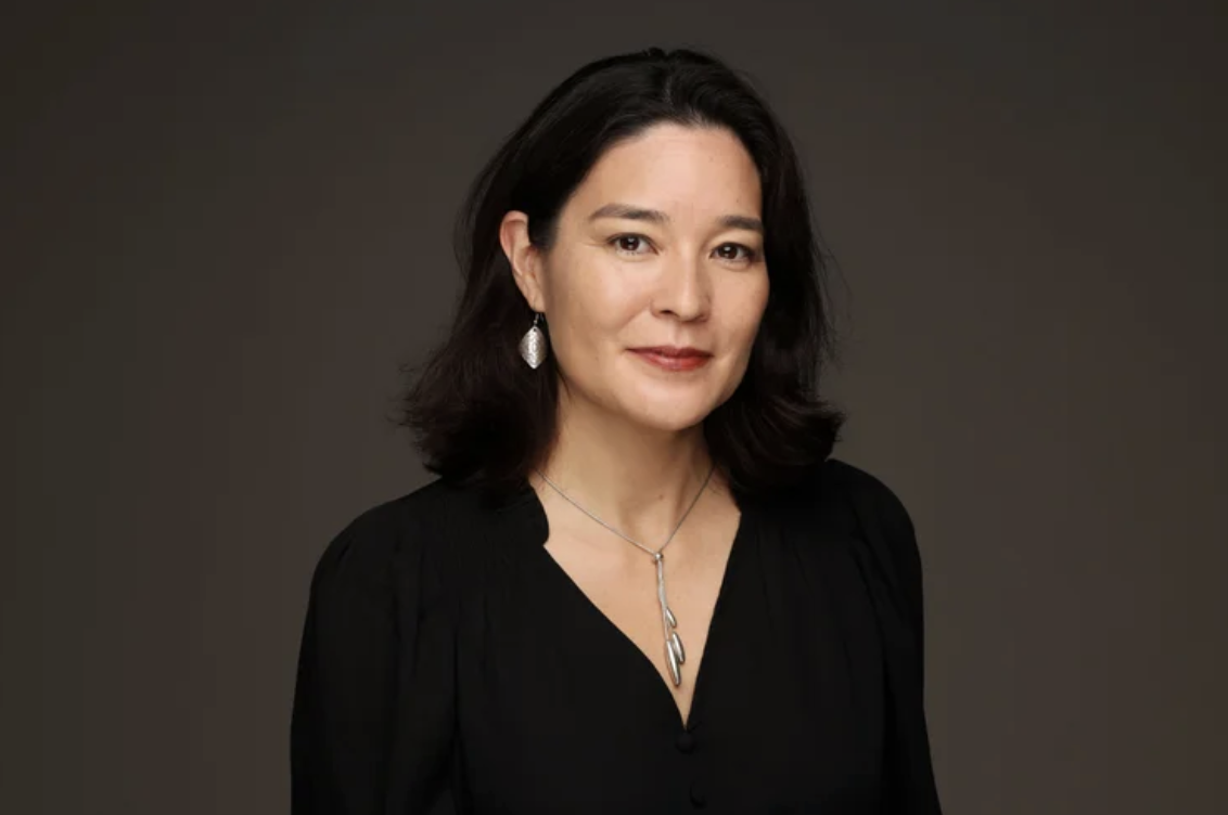 Portrait of a woman with shoulder-length dark hair wearing a black top, silver earrings, and a silver necklace against a dark background.