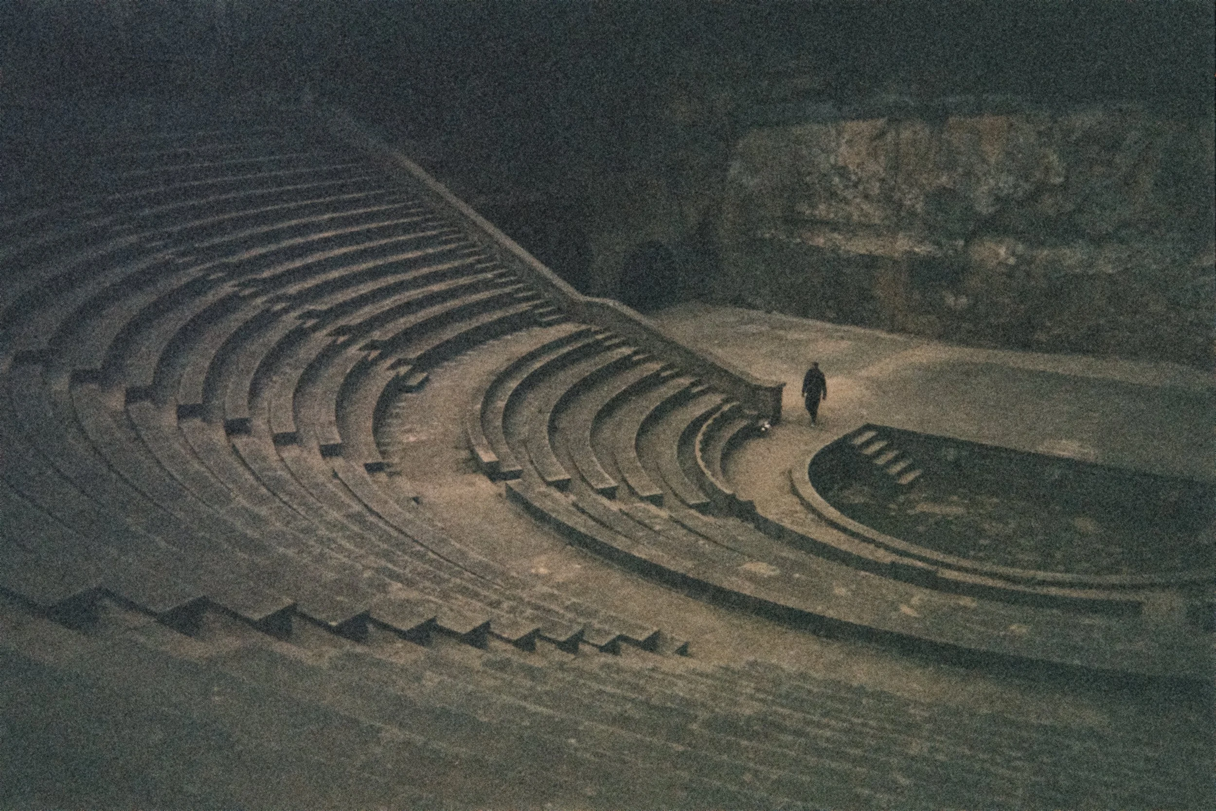 Nighttime view of an outdoor amphitheater with curved tiered seating and a small figure walking along the path.