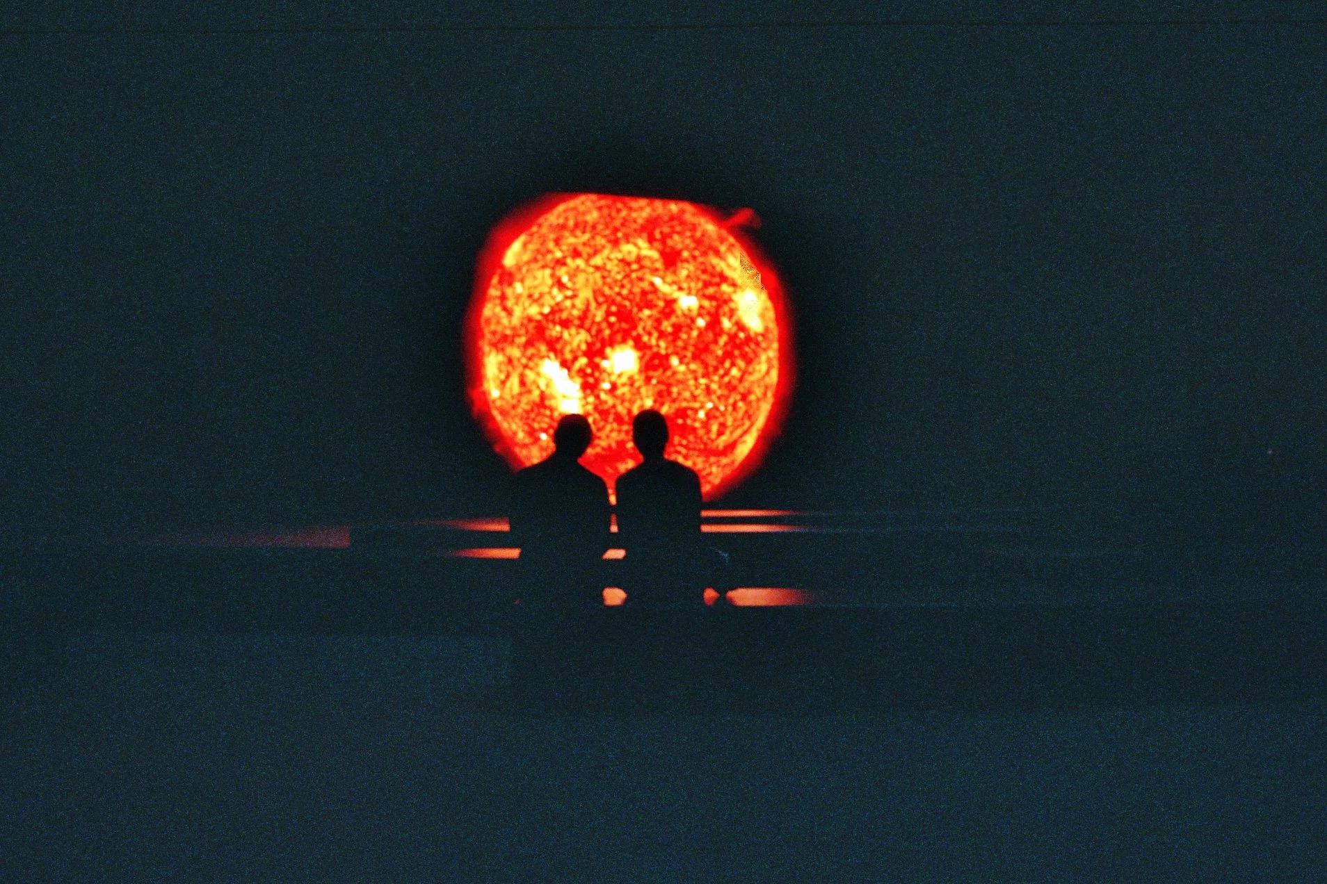 Two people sitting in front of a massive, fiery sun in space, silhouetted against the bright, glowing surface.