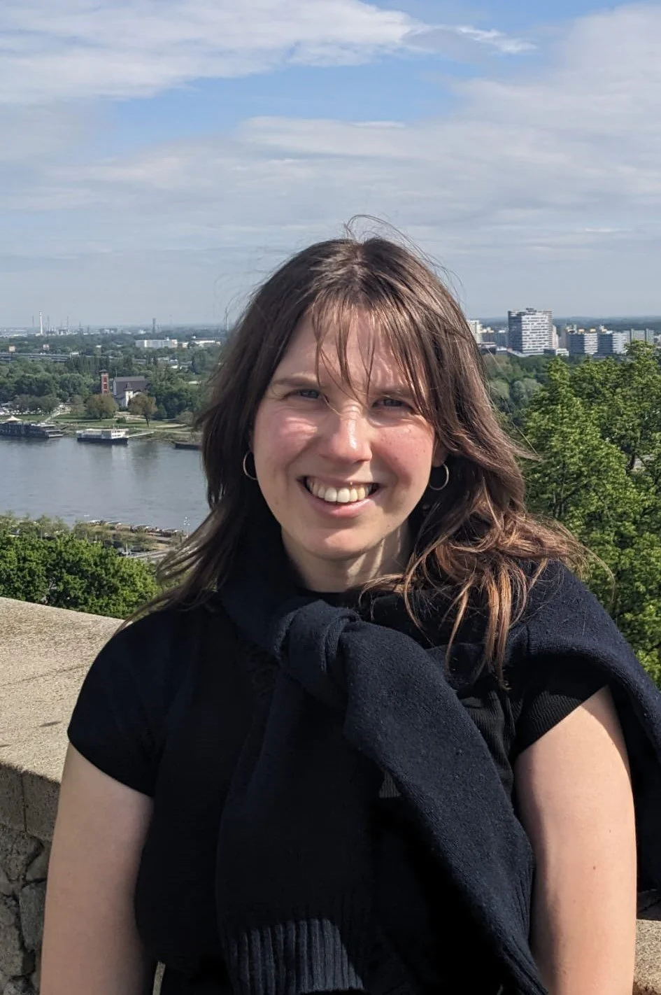 Grace Harley smiling. She has shoulder-length brown hair, wearing a black shirt and a black sweater draped over her shoulders, standing outdoors with a cityscape, river, and partly cloudy sky in the background.
