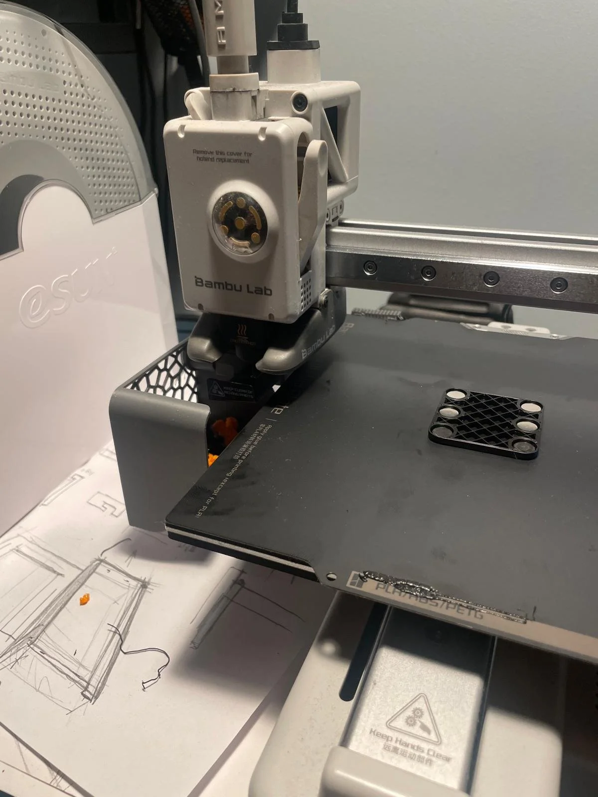 Close-up of a Bambuc Lab 3D printer extruder head during printing, with a black print bed and a black plastic component nearby, along with sketches on paper underneath.