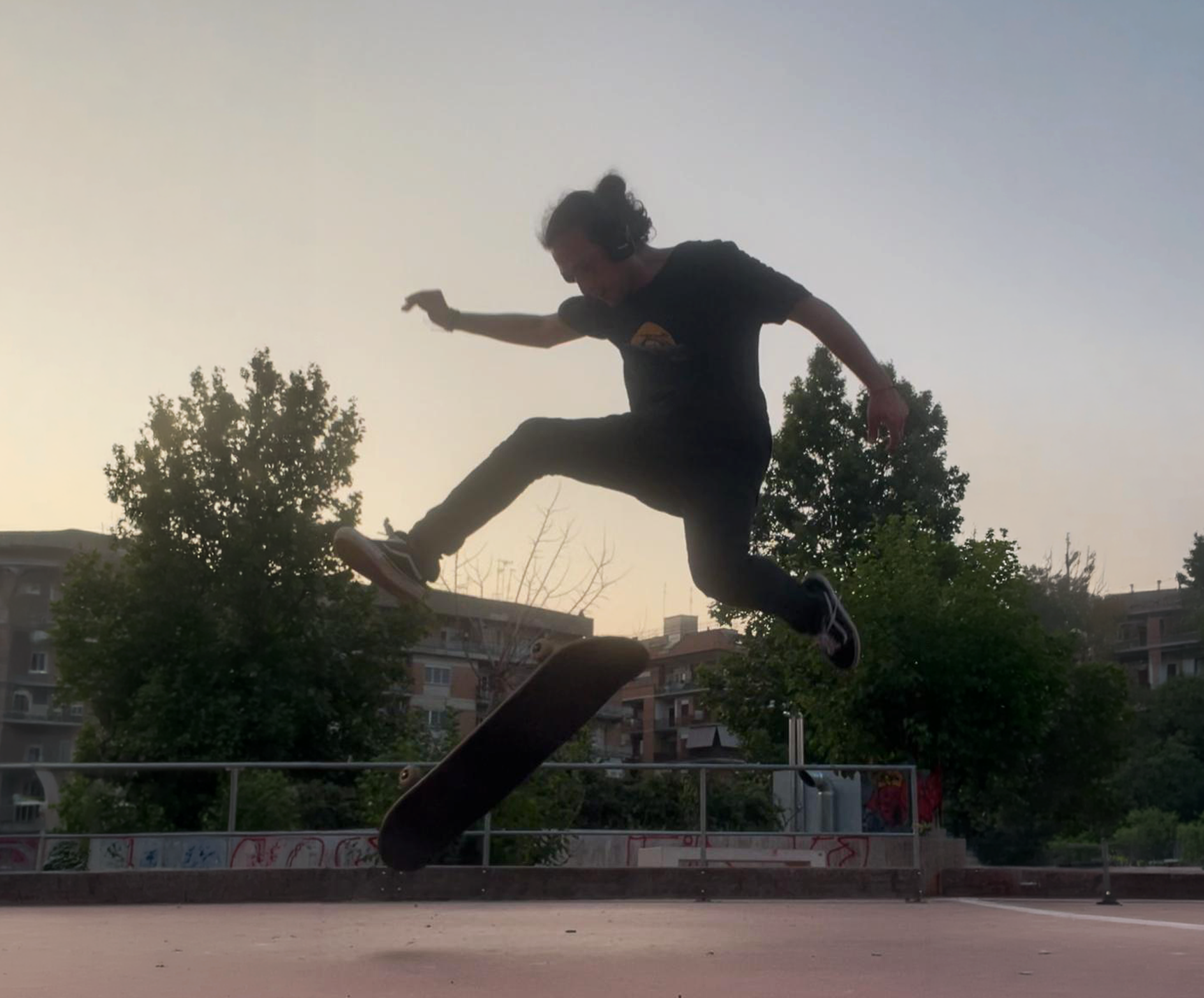 Person performing a skateboard trick in an outdoor skating area during dusk, with trees and residential buildings in the background.