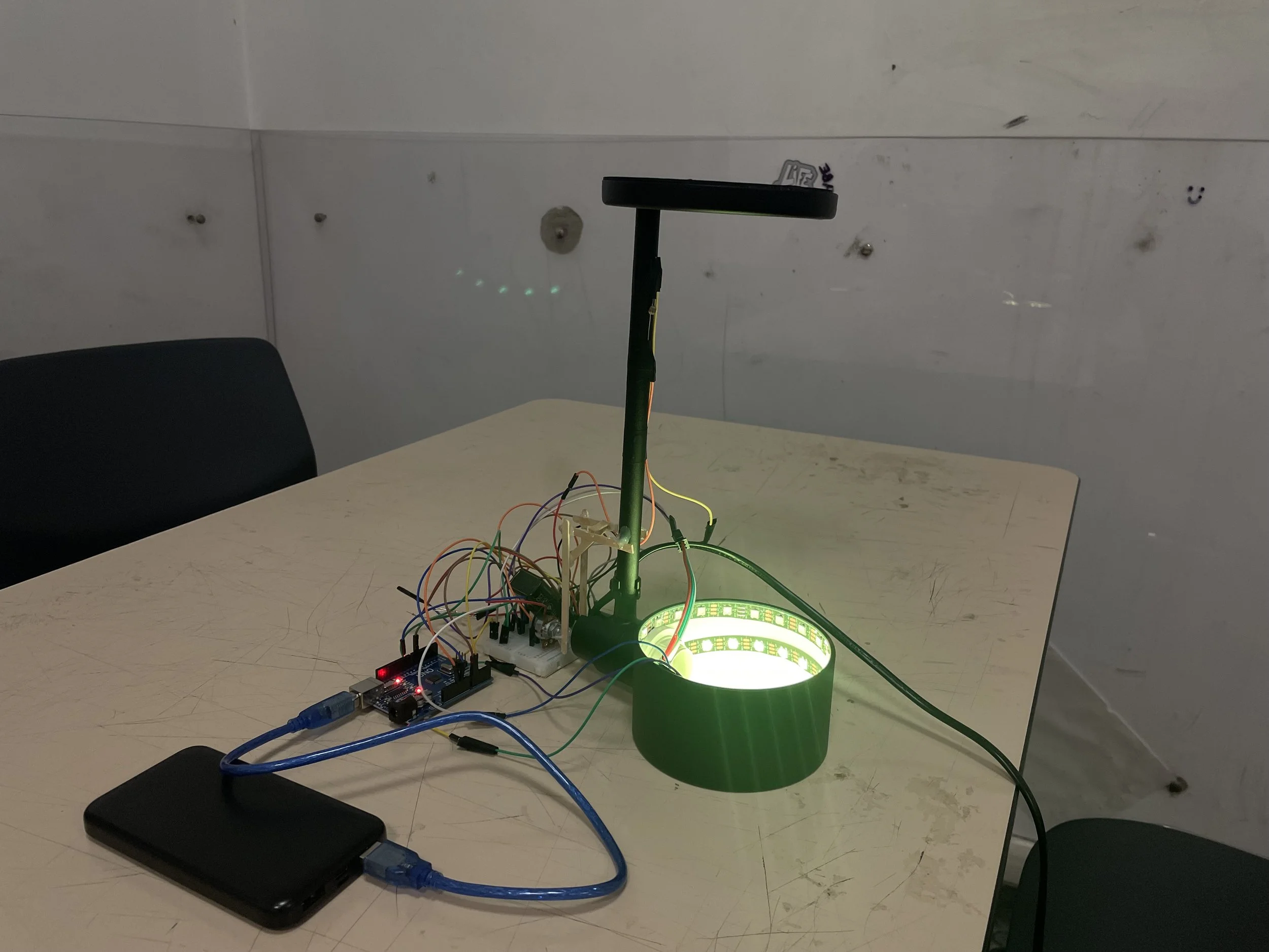 A small electronic setup on a table with a green lighted ring and wires connected to a microcontroller, a U-shaped stand above the setup, in a room with white walls and two black chairs. An arduino project.