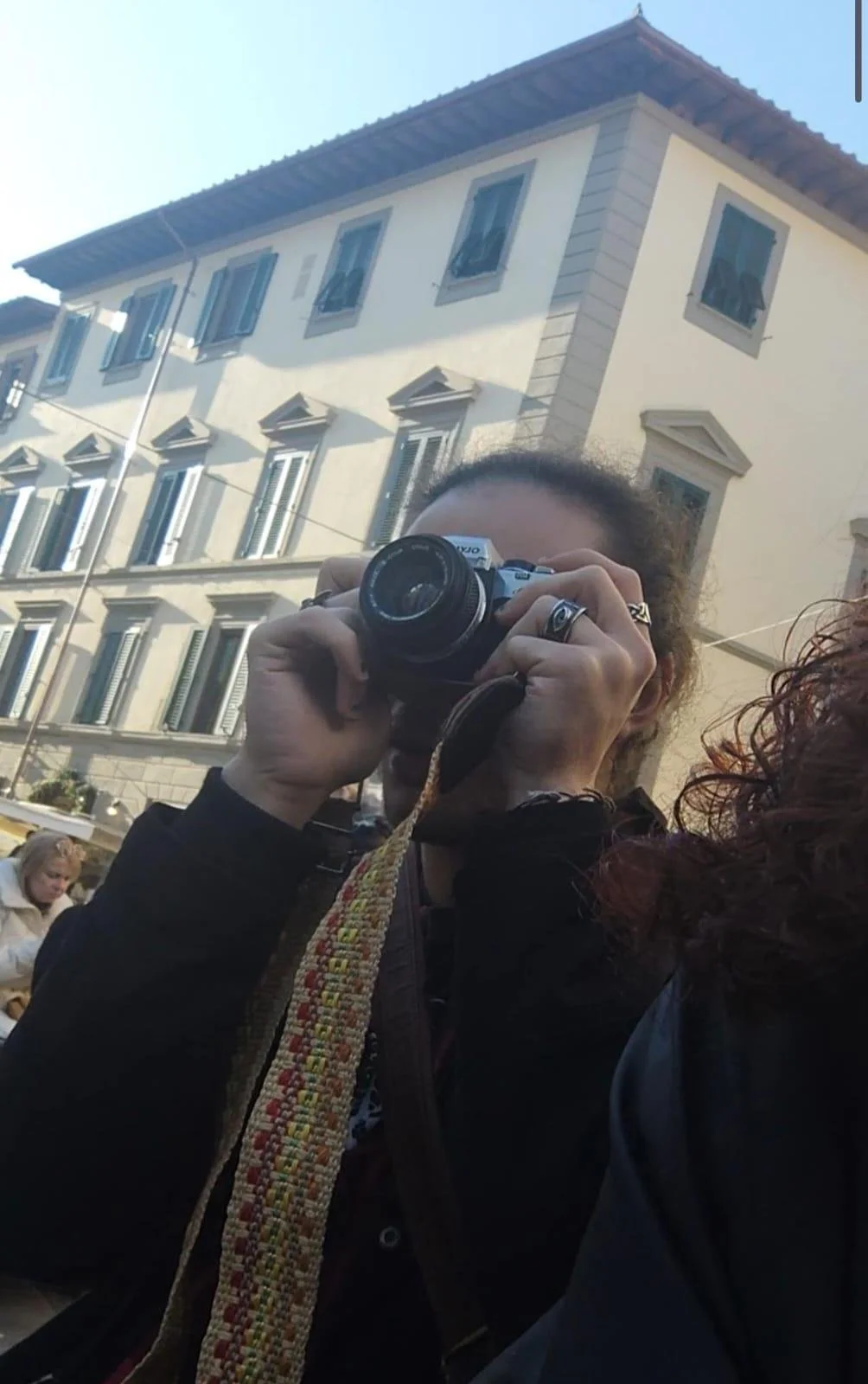 Person with curly hair taking a photo with a camera on a city street, buildings with multiple windows in the background.