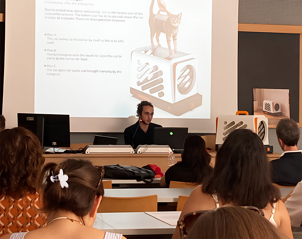 A presenter explains a project about a cat and a sorter machine to an audience in a classroom or conference room. The presentation slide shows an illustration of a cat on top of a machine with a slot, possibly for food, along with text describing different plans for the machine.