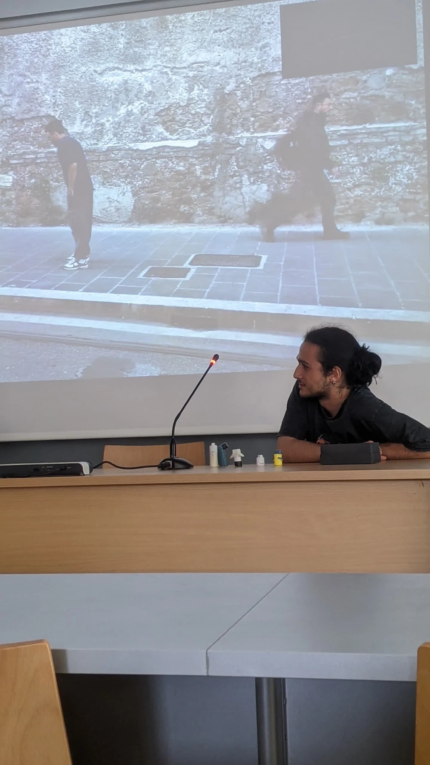 A man with dark curly hair tied in a bun sits at a desk in a classroom or conference room, with a large projection screen behind him. The projected image on the screen shows two people walking by a stone wall, one with a backpack.