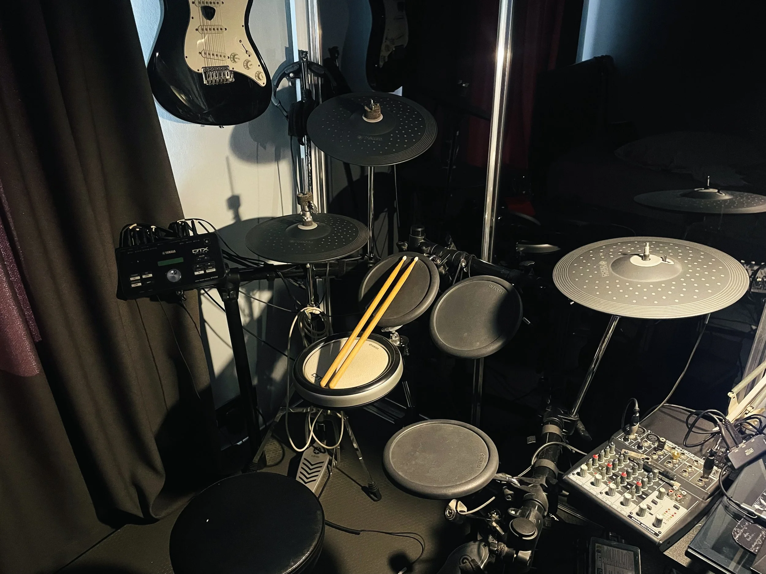 A drum set and electric guitar on a stand with a sound mixer and headphones nearby, in a dark room, possibly a recording studio. Yamaha DTX 532K.