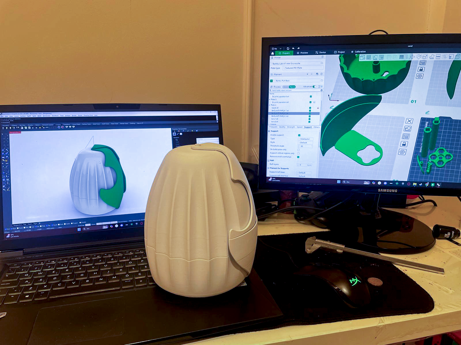 A workspace with two computer monitors displaying 3D printing software, a white 3D printed object in front of a laptop.