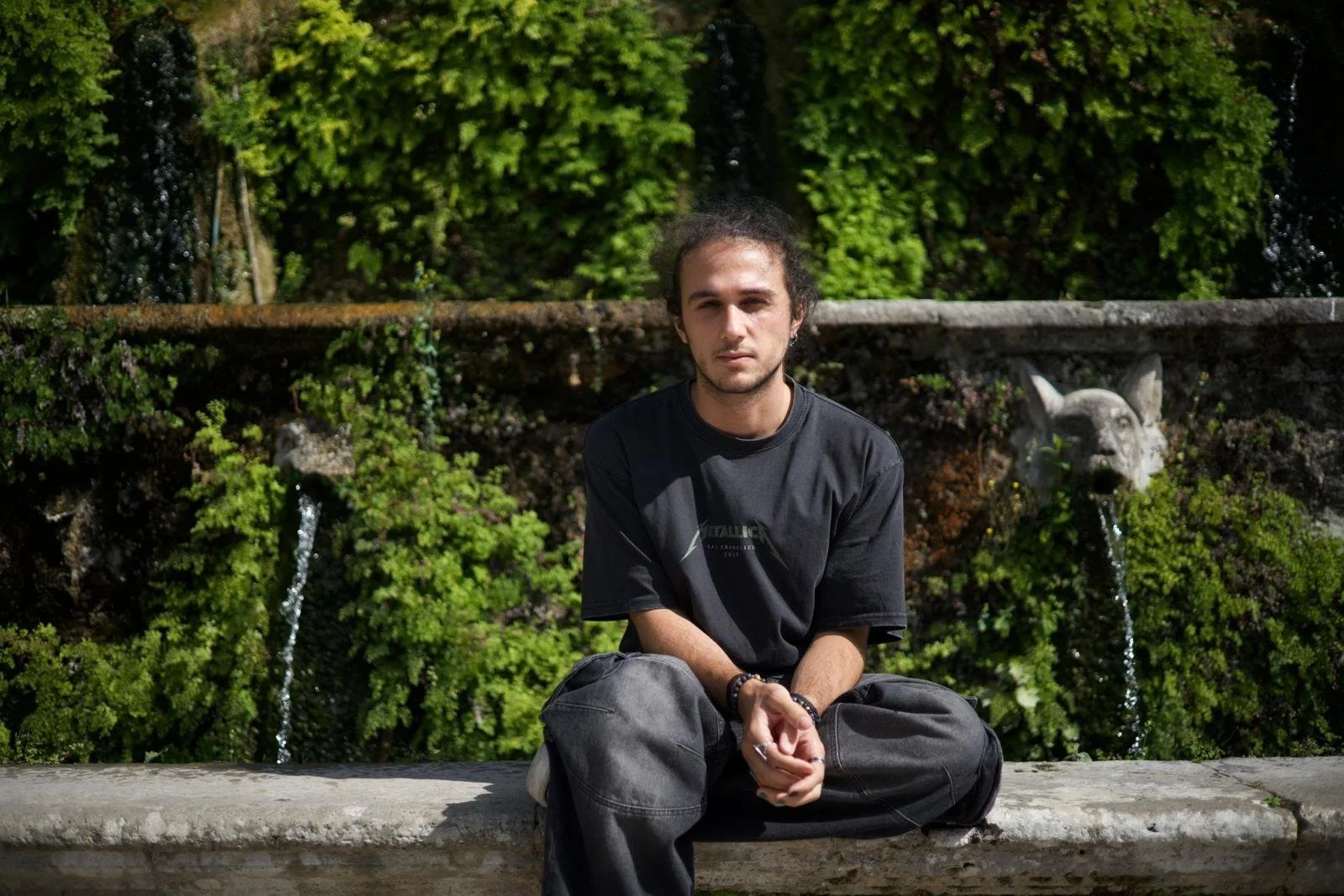 A young man with dark, curly hair and light skin sitting on a stone bench in front of a green shrub wall with a fountain featuring a lion's head spouting water.