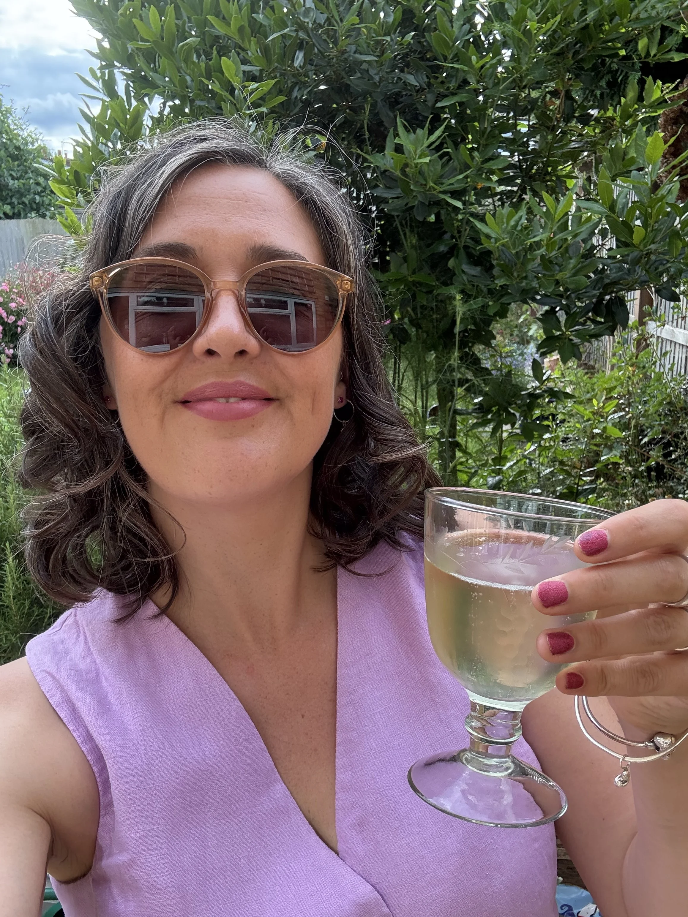 A woman with curly gray hair, wearing sunglasses and a light purple sleeveless top, holding a glass of white wine outdoors with green foliage in the background.