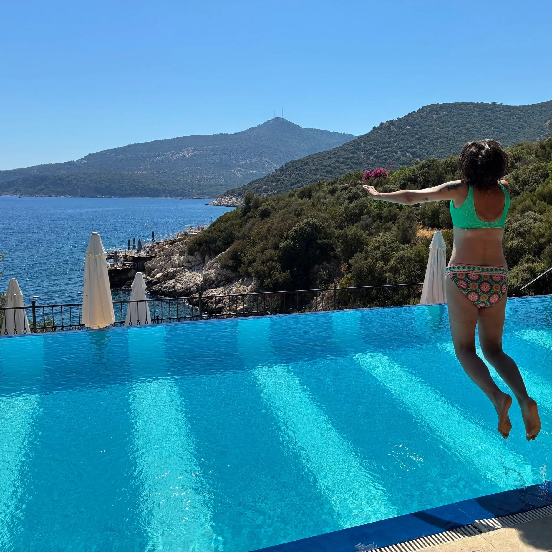 Woman in a green bikini standing by a swimming pool with arms outstretched, overlooking a mountainous landscape with water and a clear blue sky.
