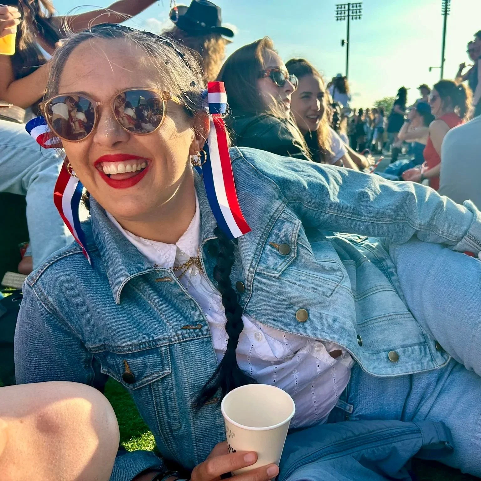 A woman wearing sunglasses with patriotic ribbons in her hair, smiling at an outdoor event, holding a paper cup. There are other people sitting on the grass in the background.
