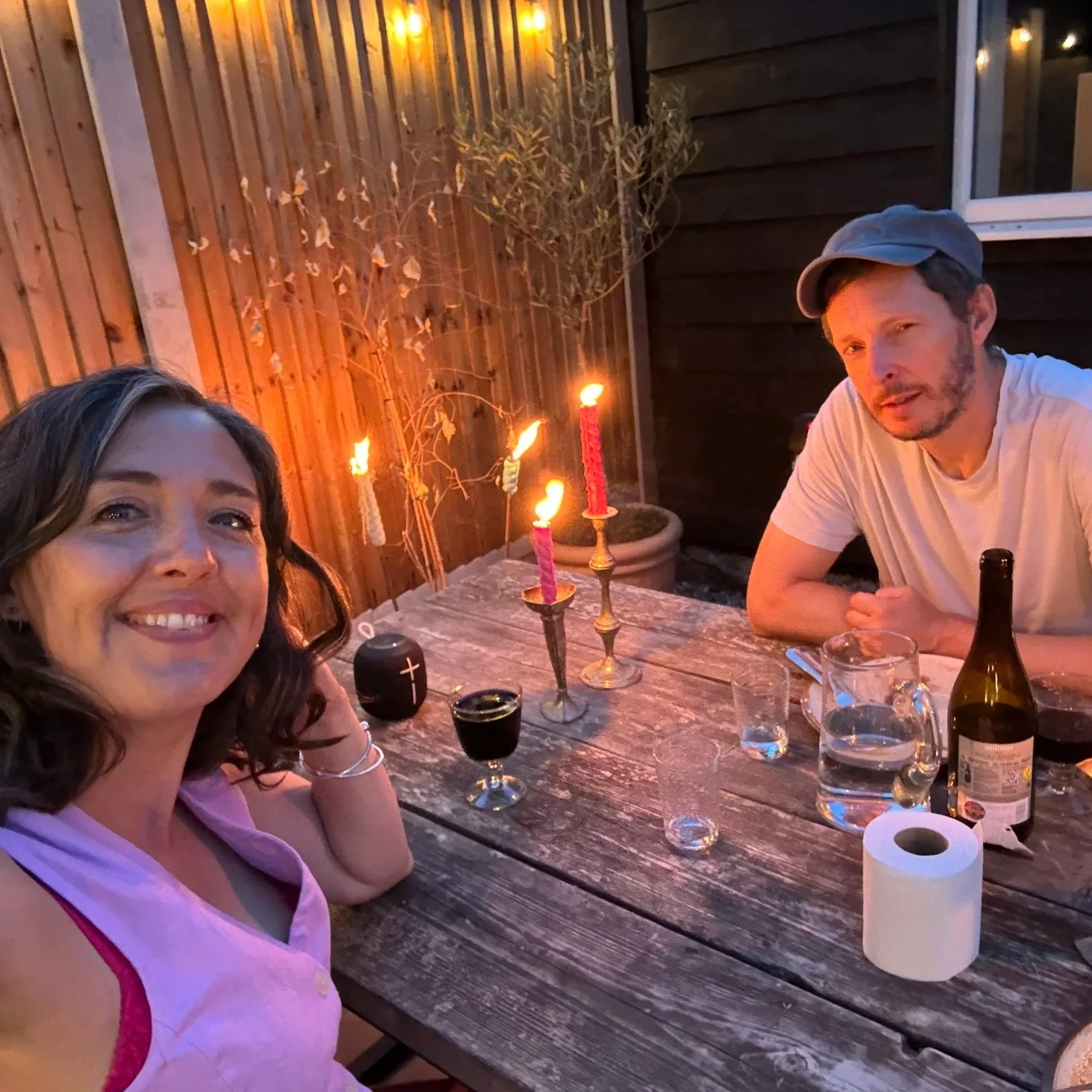 A woman with brown hair smiling at a dinner table with a man with a beard and cap; the table has candles, water bottles, and drinks, with a wooden fence and some outdoor lights in the background.