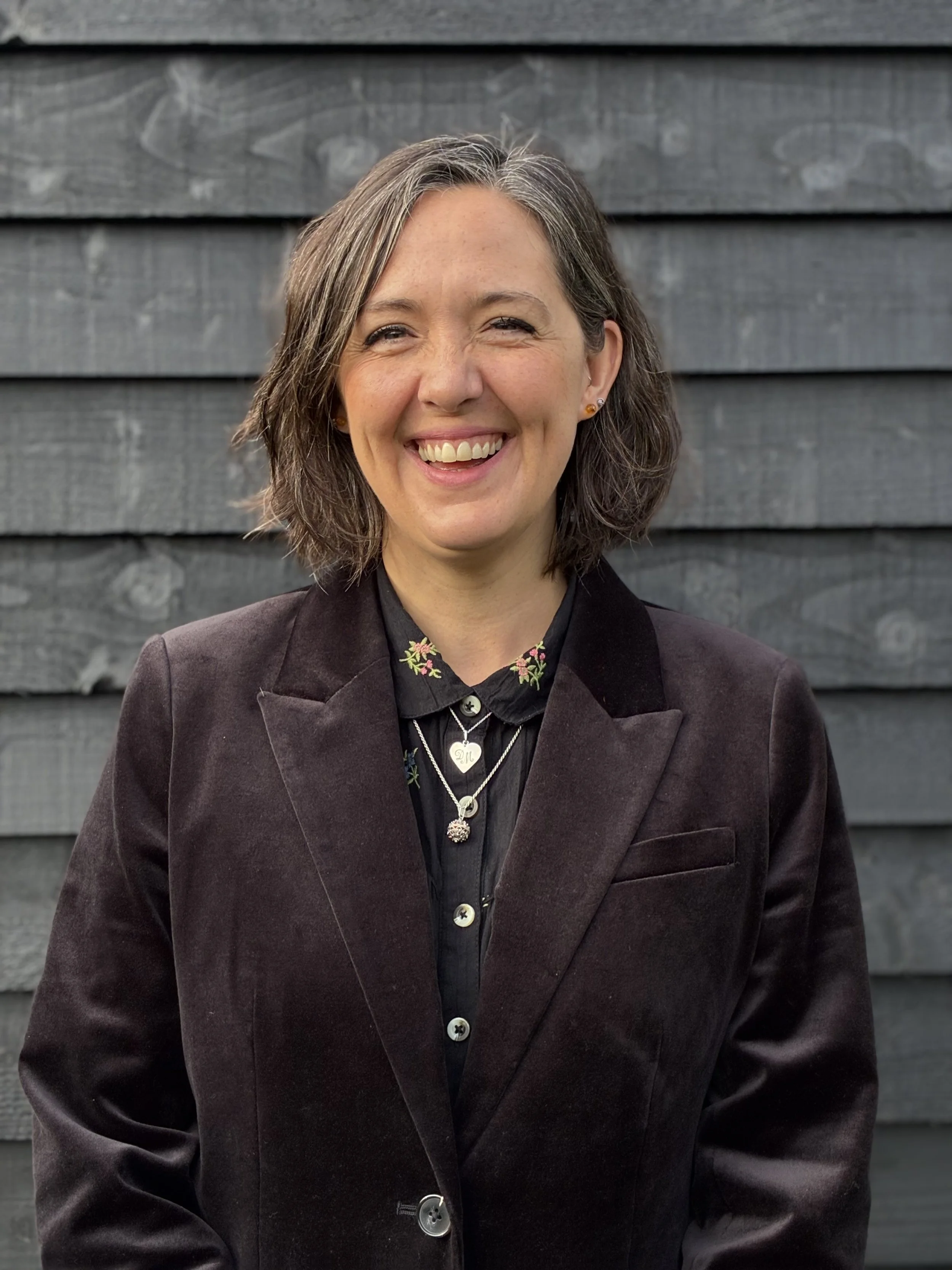 A woman with shoulder-length gray hair, smiling, wearing a black collared shirt with floral embroidery, layered necklaces with heart and spherical pendants, and a dark blazer, standing in front of a wooden wall with horizontal gray planks.