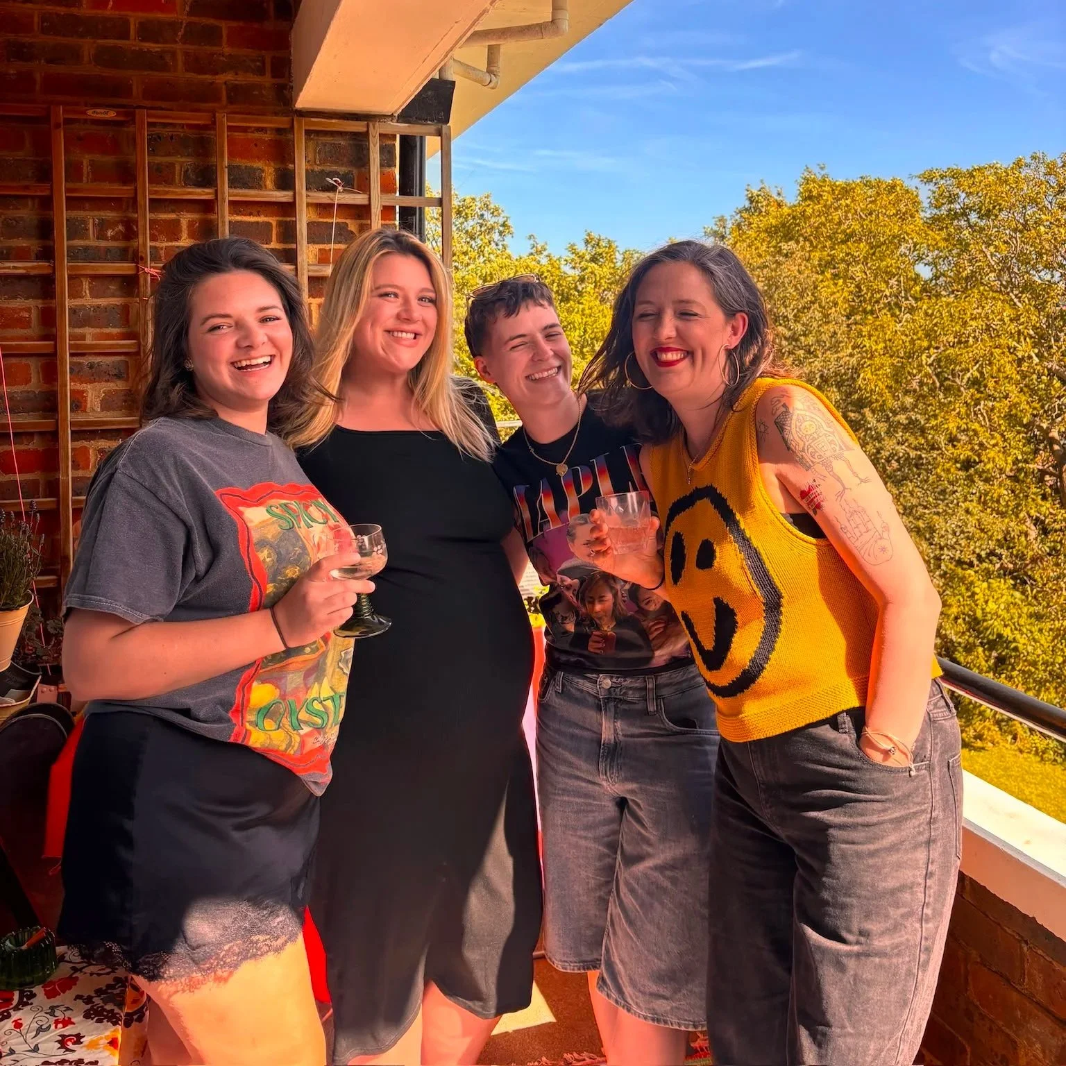 Four women smiling and holding drinks on a balcony with trees and a blue sky in the background.