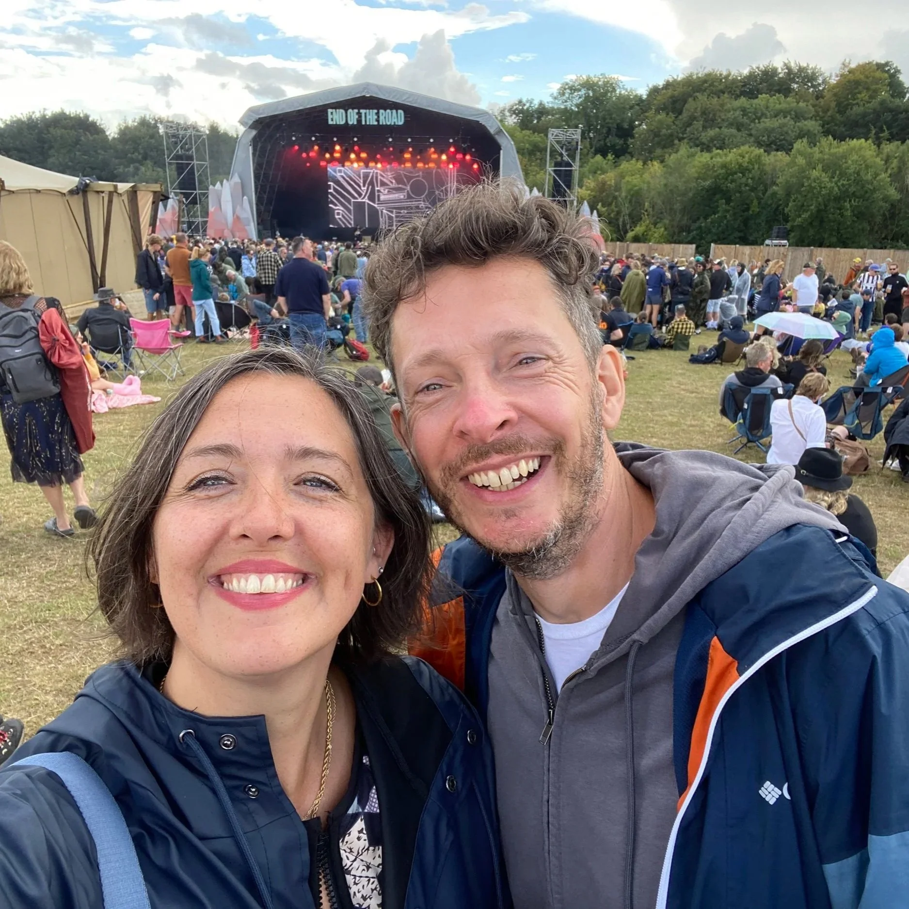 Two people smiling for a selfie at an outdoor music festival with a crowd and stage in the background. The stage has a sign that reads "End of the Road."