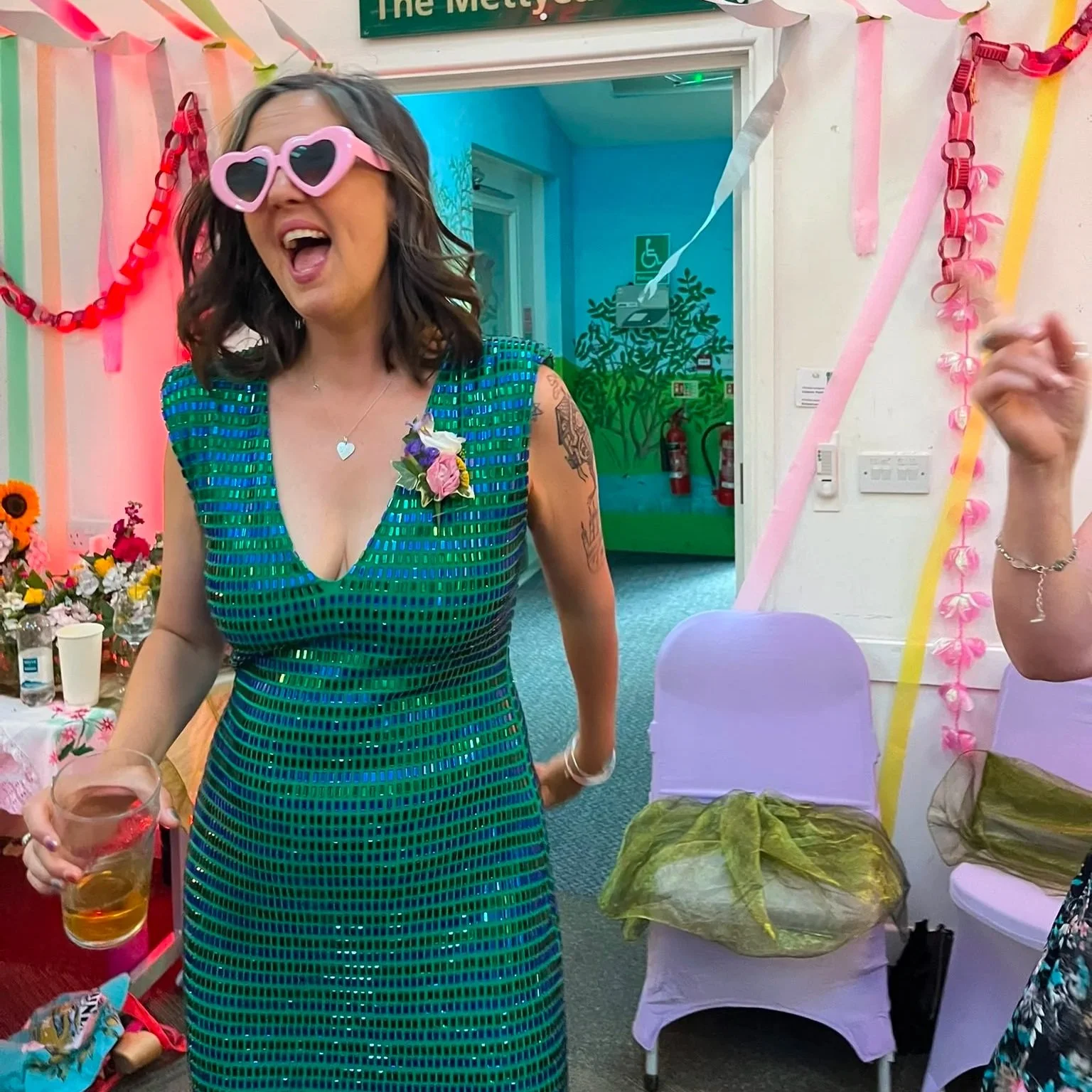 Woman wearing heart-shaped sunglasses and a green dress with blue vertical stripes, smiling and holding a drink at a party decorated with pink, yellow, and green streamers and flowers.