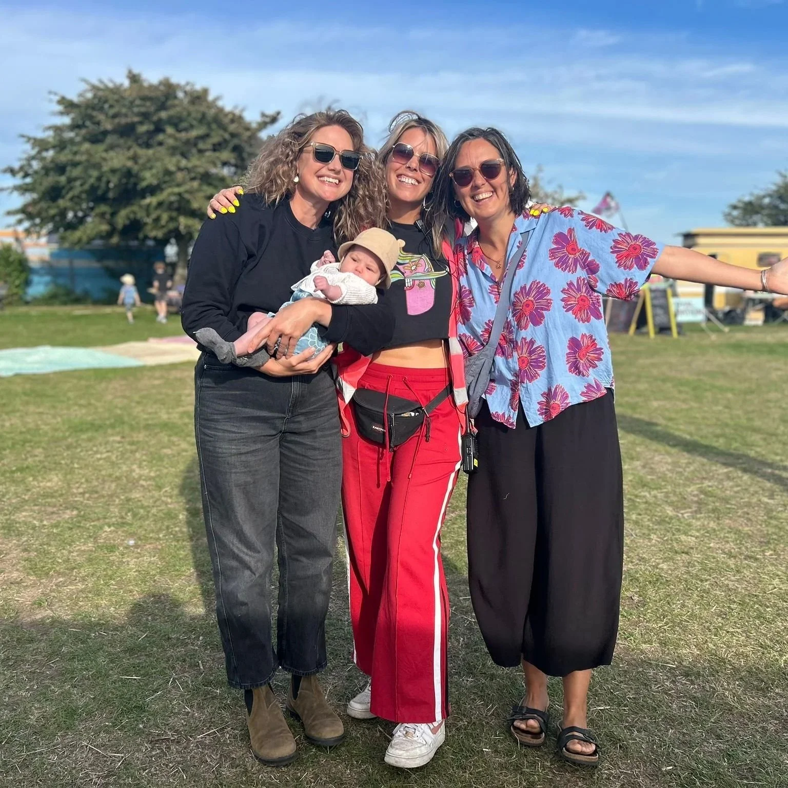 Three women and a baby standing outdoors on a grassy field, smiling and posing for the camera with a tree and a clear blue sky in the background.