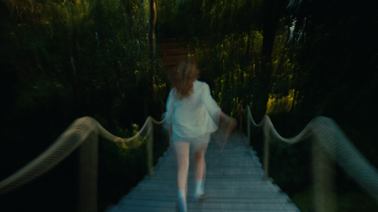 A woman with curly hair walking down a wooden bridge in a dense forest during daytime.