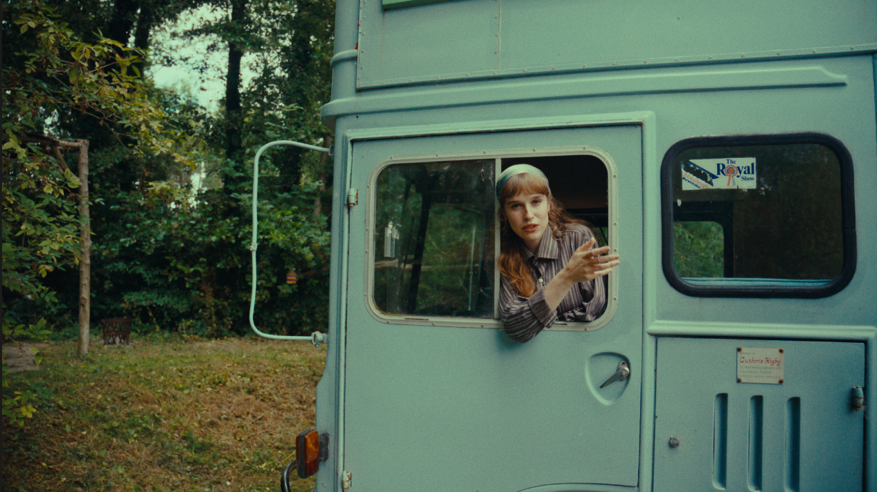 A woman with reddish hair, wearing a striped shirt and a headband, leans out of a light blue food truck or camper, gesturing with her hand, outdoors with trees and greenery in the background.
