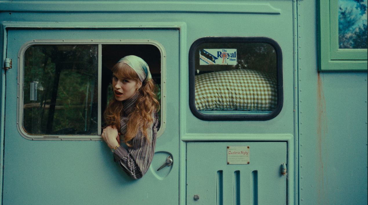 A woman with red hair and a headscarf looking out of the window of a light blue camper trailer.