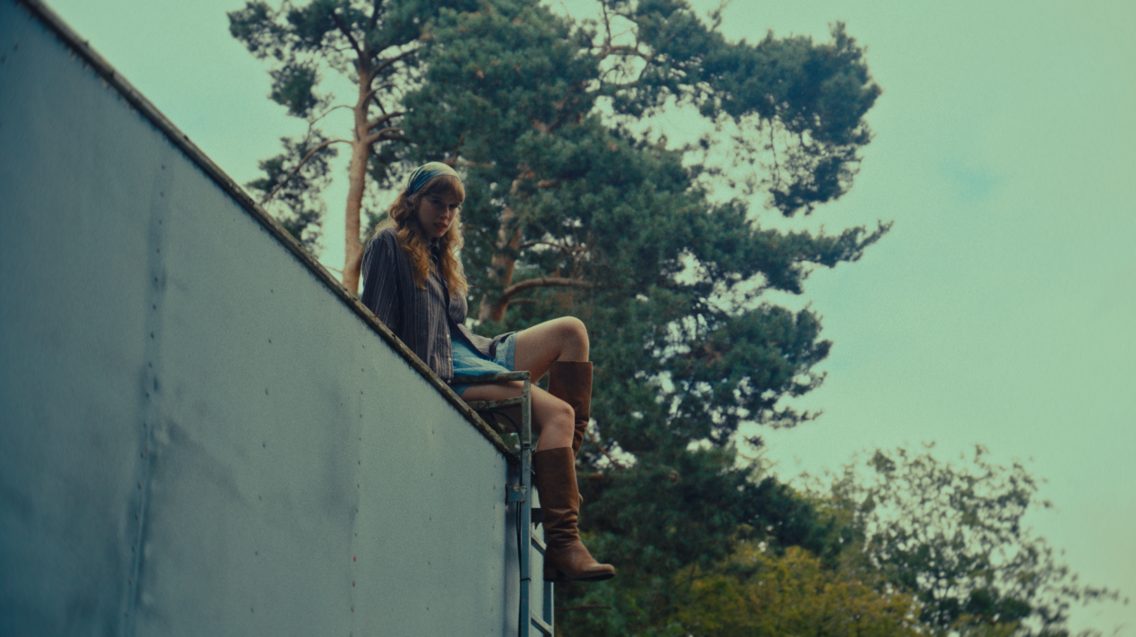 A young woman sitting on the edge of a rooftop, looking down with trees in the background.