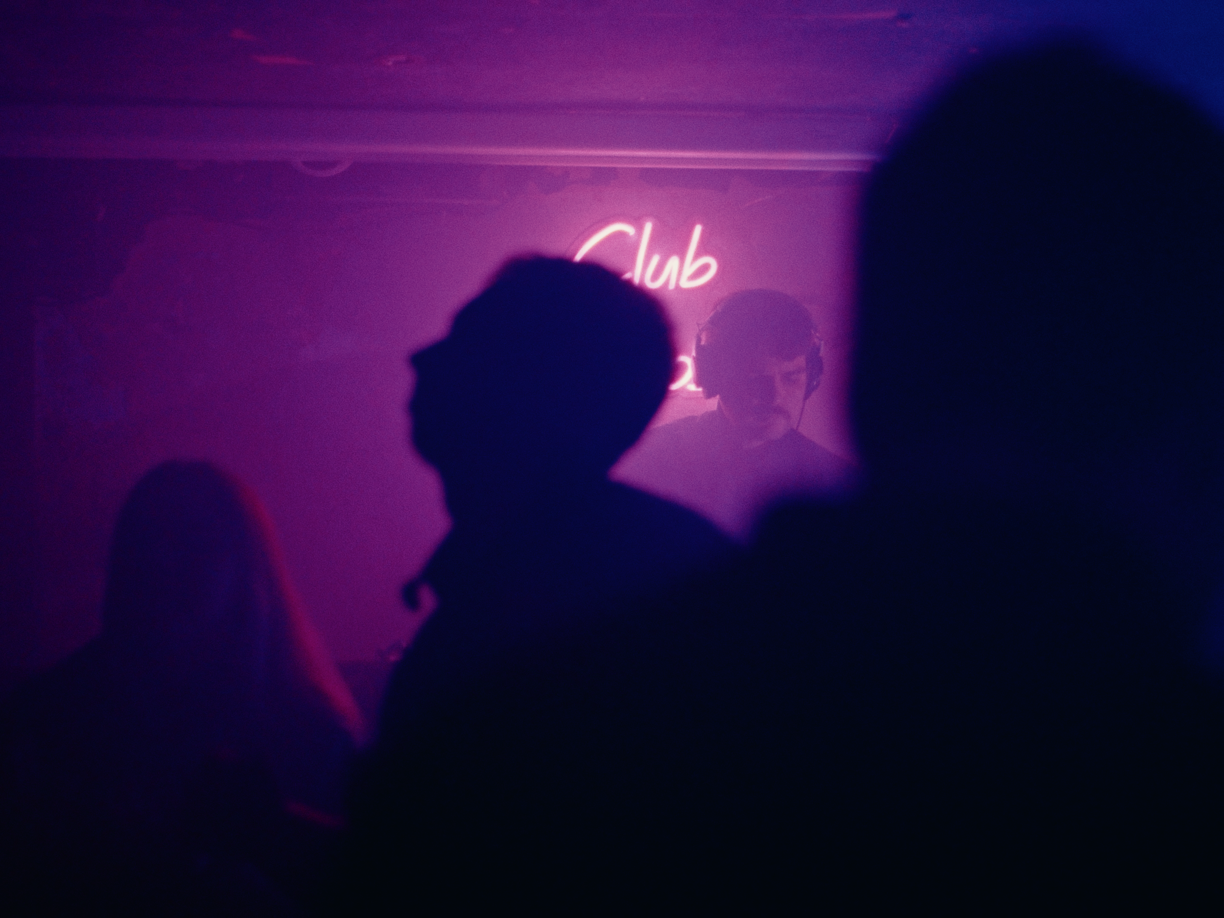 A dimly lit nightclub scene with silhouettes of people dancing and a neon sign that says 'Club' in pink. A DJ wearing headphones is visible in the background.