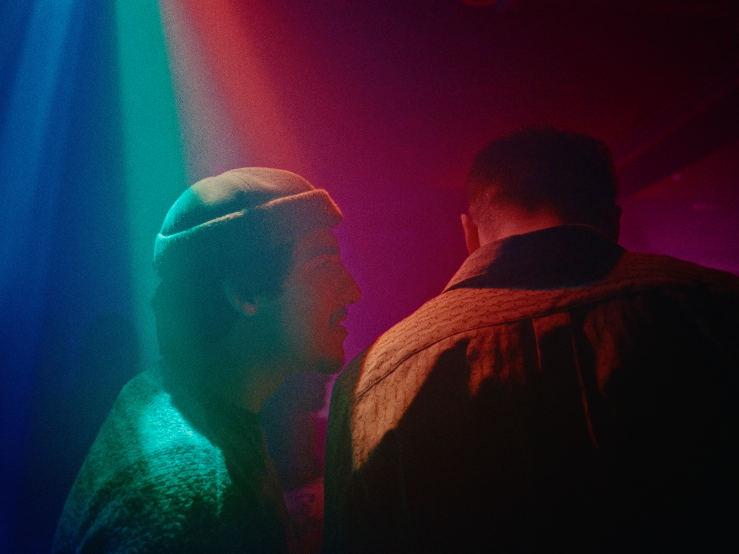 Two young men having a conversation in a dimly lit space with colorful lighting effects.