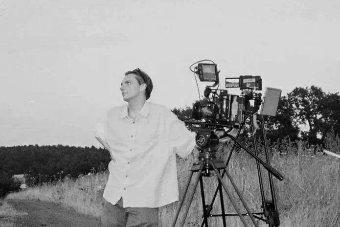 A man in a white shirt standing outdoors next to a professional film camera on a tripod, with a landscape of trees and a dirt path in the background.