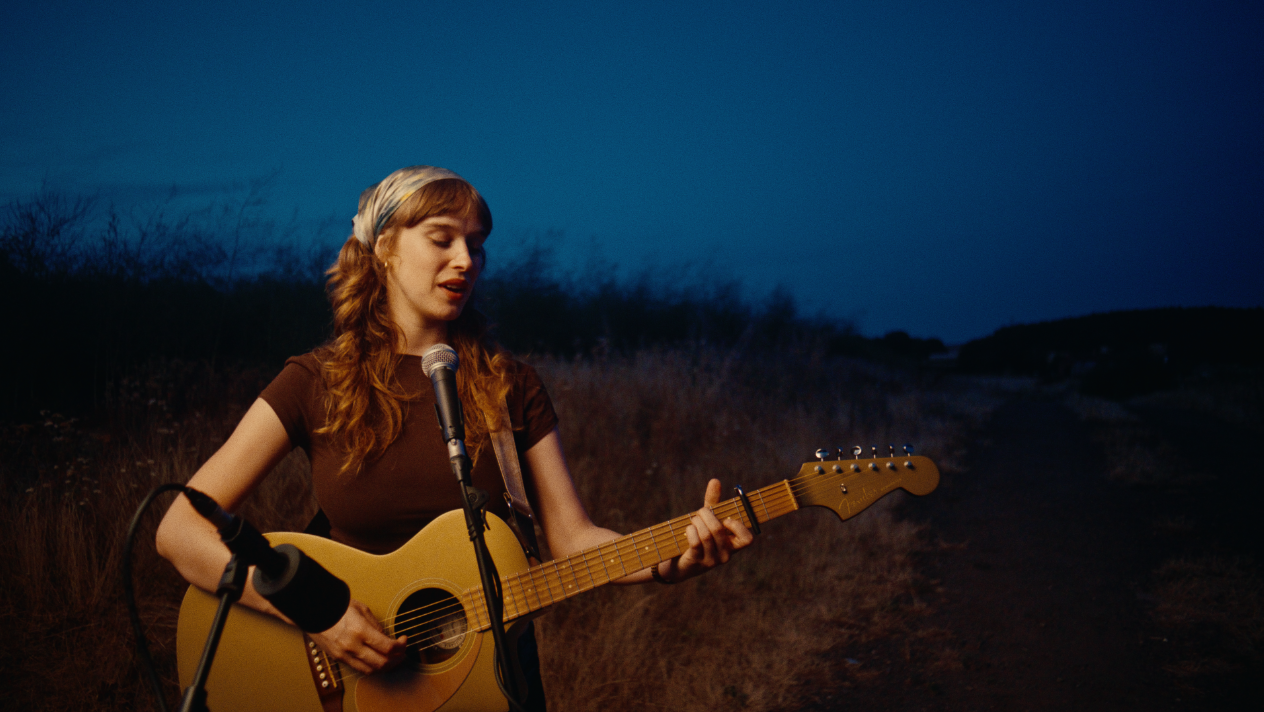 A young woman with long, wavy hair playing an acoustic guitar and singing into a microphone outdoors during twilight.