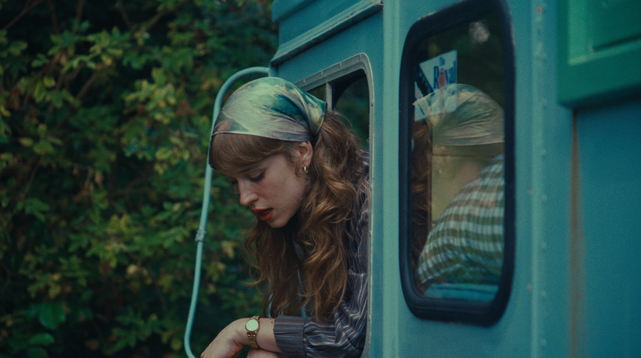 A woman looking down, leaning out of a vintage camper trailer window, with greenery in the background.