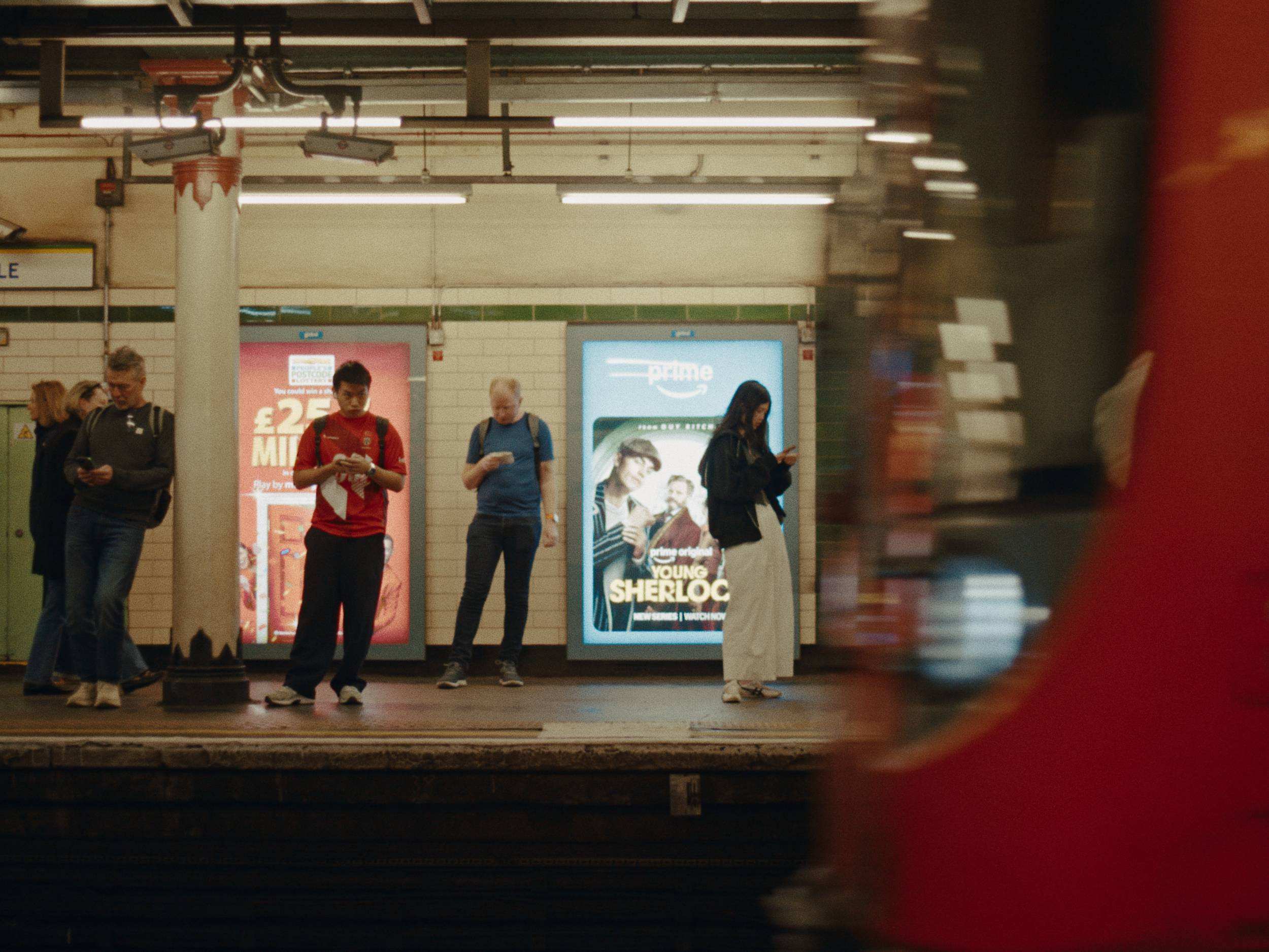 People standing at the subway station looking at their phones, with a brightly lit advertisement in the background, and a blurred person moving in the foreground.