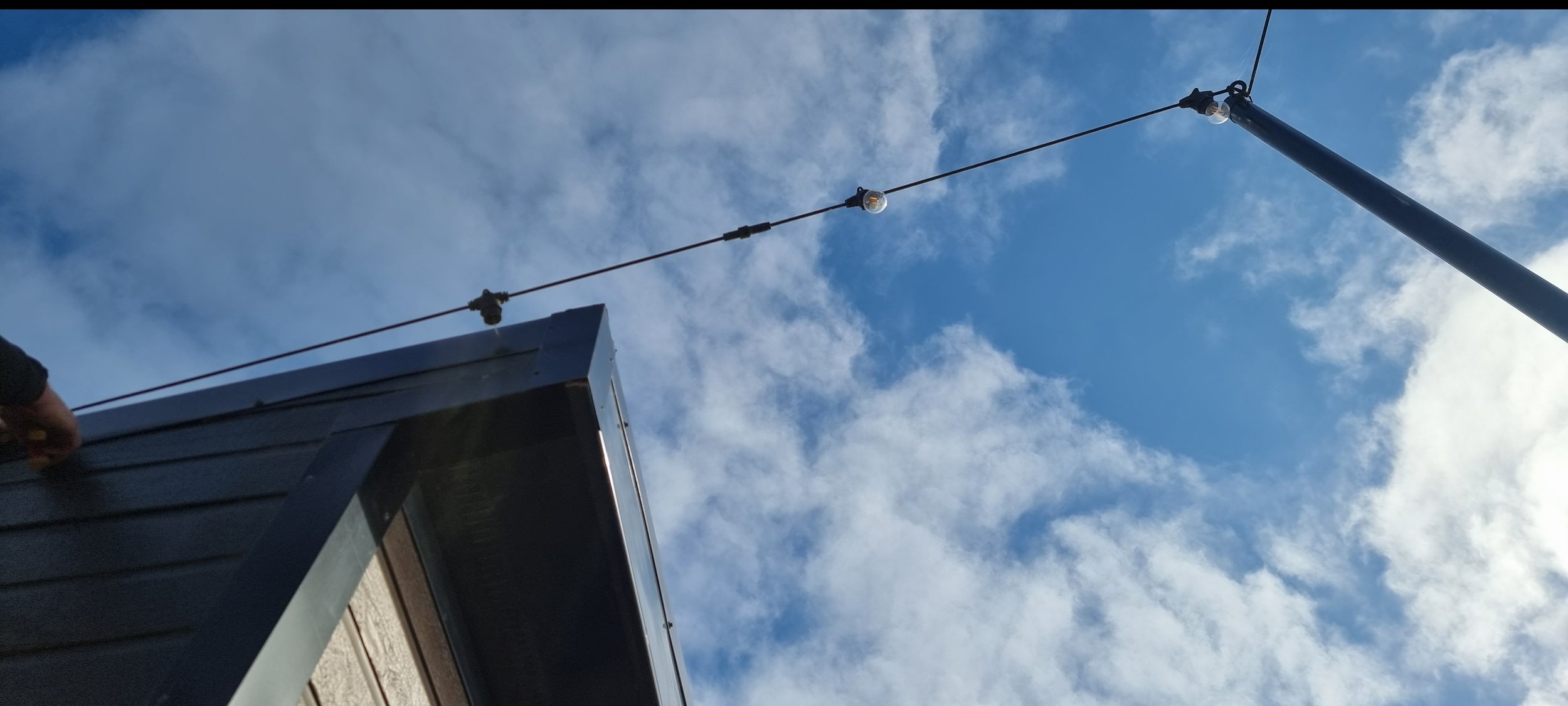 View of a rooftop with a metal railing and a tall antenna or pole attached to the side, set against a partly cloudy sky.